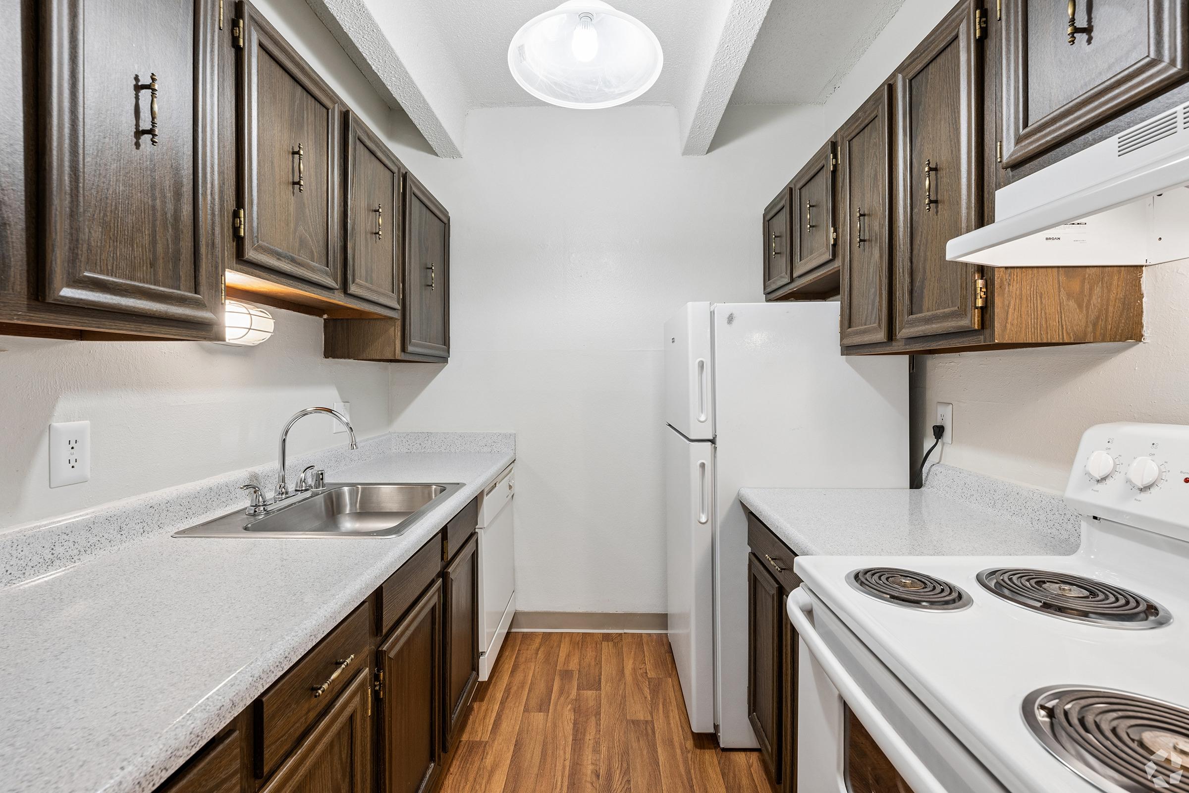 A small kitchen featuring dark wooden cabinets, a double sink, a white refrigerator, and a white stove with an oven. The countertops are light gray, and the flooring is laminate wood. A ceiling light fixture provides illumination, and the walls are painted a neutral color.