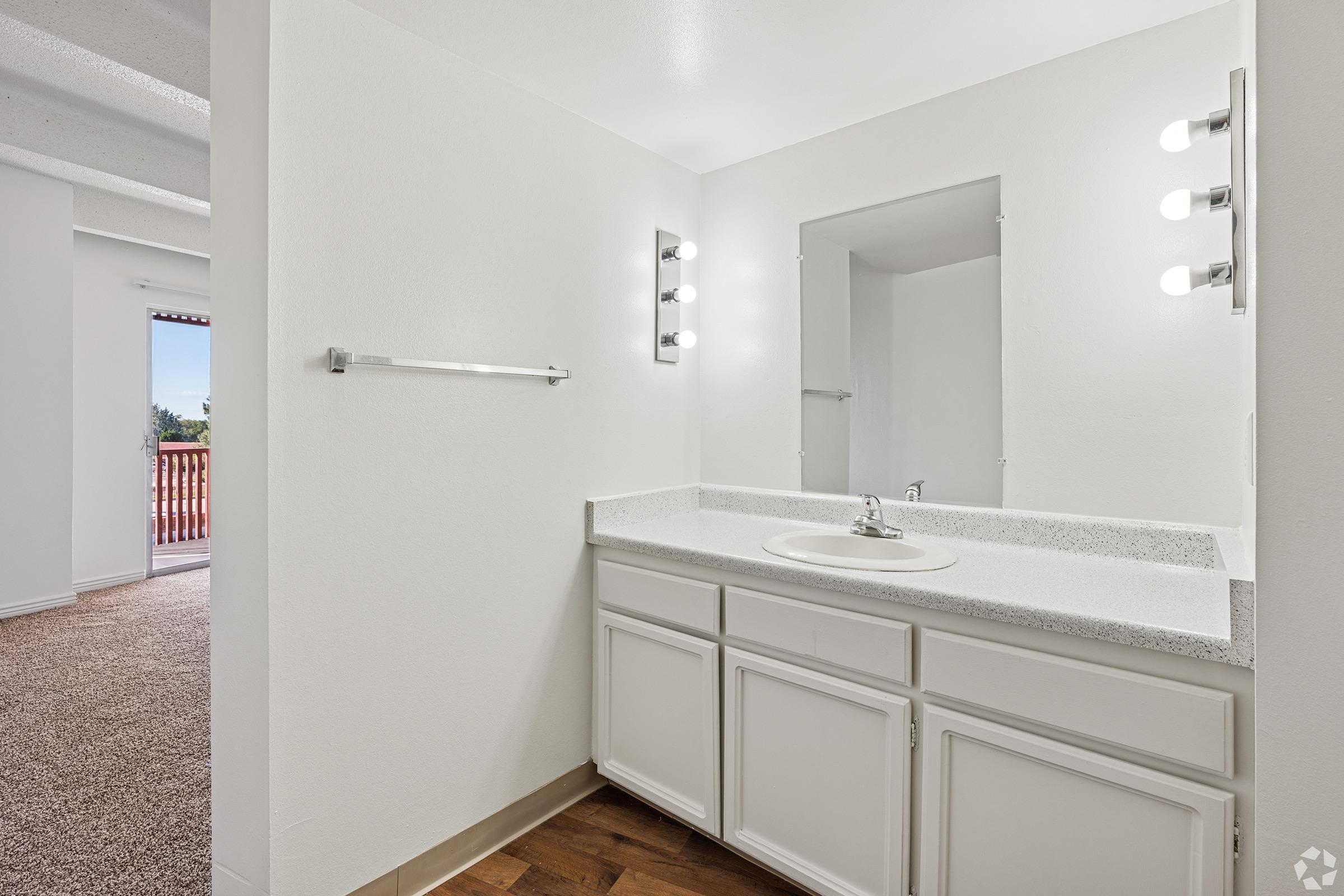 A modern bathroom featuring a white vanity with a sink, a large mirror, and bright lighting. The walls are painted white, and the floor is a warm-toned wood. A doorway leads to another room, which is partially visible, and there is a towel bar on the side wall.