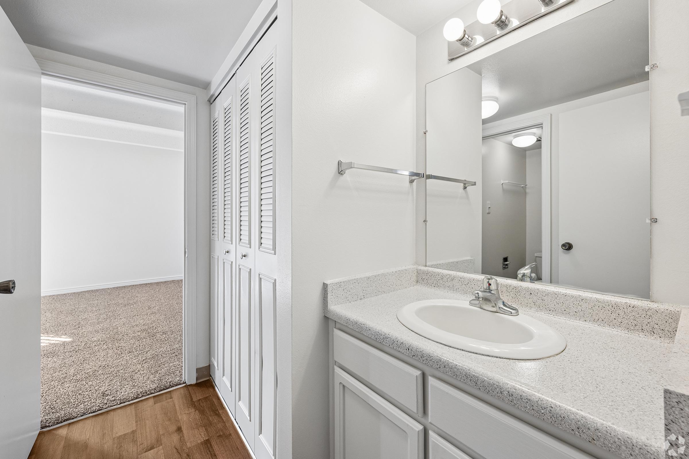 A modern bathroom featuring a single sink with a faucet on a granite countertop, a mirror above, and a white cabinet below. Light fixtures are mounted above the mirror. The image shows an open door leading to a carpeted room in the background, with neutral-colored walls.
