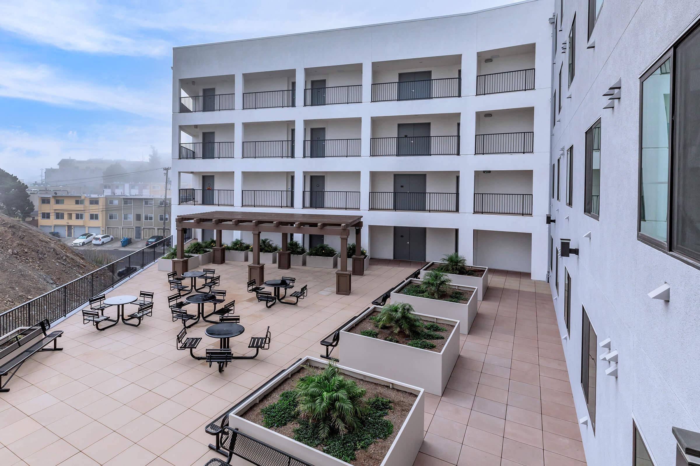 Apartments in Daly City For Rent - Brunswick Street Apartments - Outdoor Courtyard With Seating Areas and Tables, Wooden Canopy and Planters surrounded by the Building.