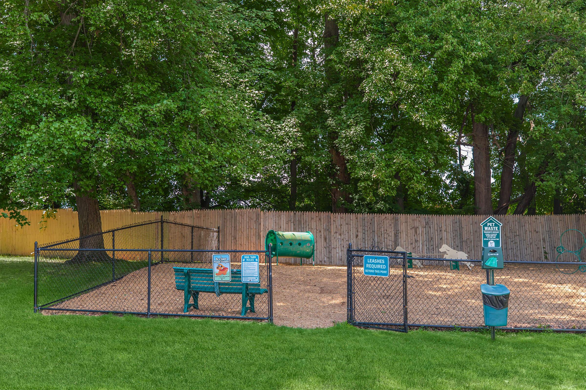 A fenced dog park featuring a grassy area with benches, a waste disposal station, and agility equipment, surrounded by tall trees and a wooden fence in the background.