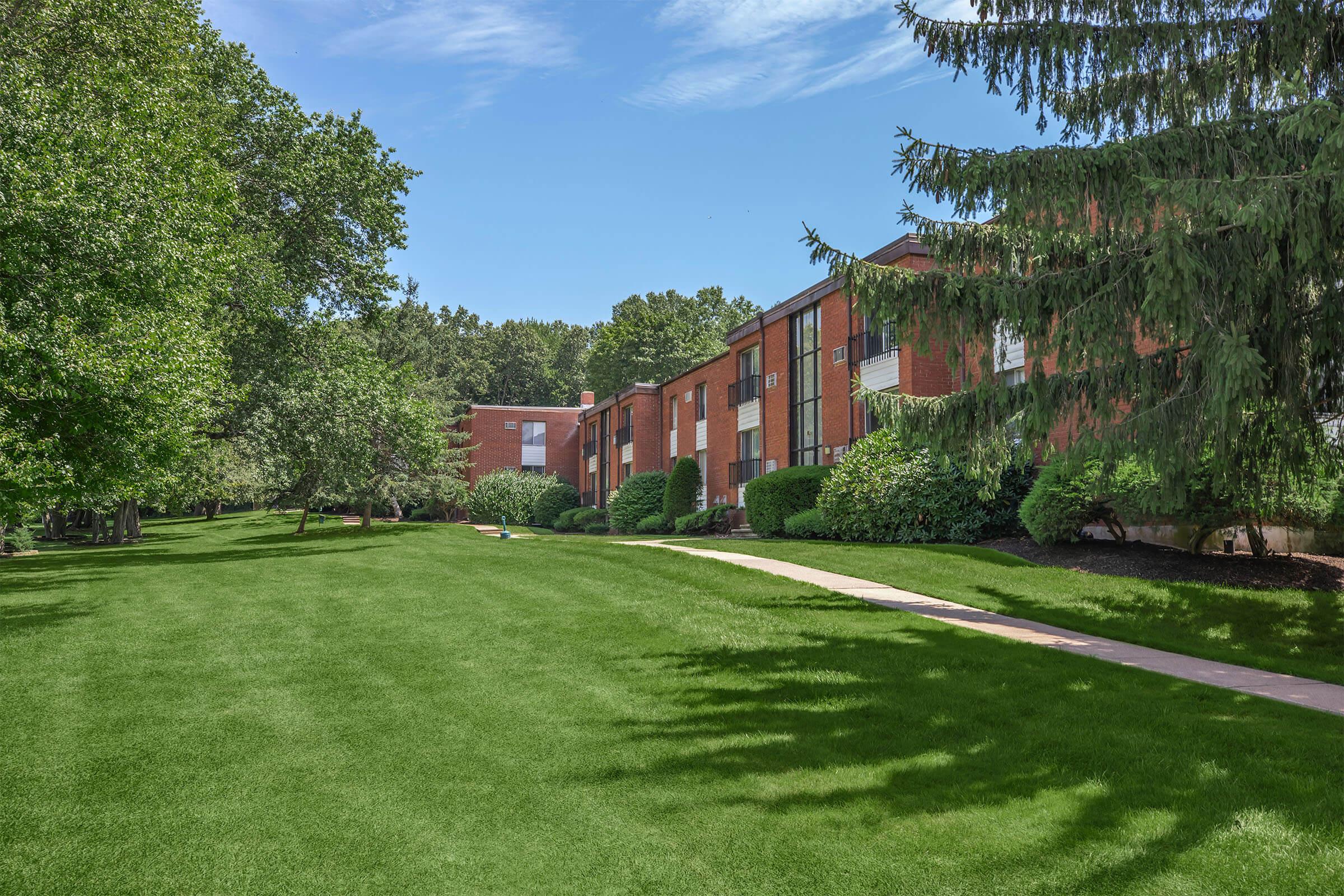 A well-maintained residential area featuring brick apartment buildings surrounded by lush green grass and trees. The pathway leads through the landscaped grounds, creating a peaceful and inviting atmosphere under a clear blue sky.