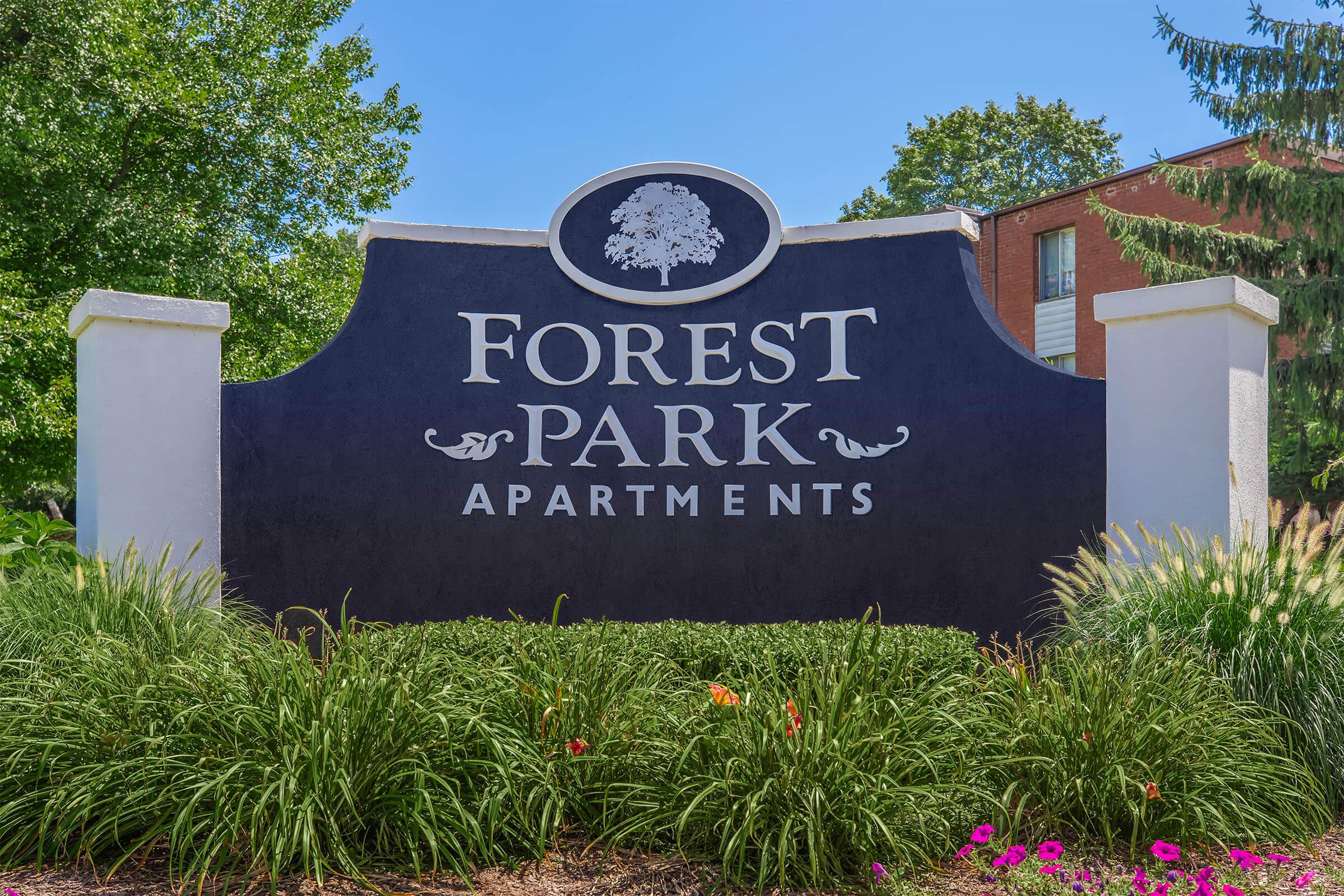 Sign for Forest Park Apartments featuring bold white lettering on a dark background. The sign is flanked by green foliage and colorful flower beds, set against a clear blue sky and trees in the background, indicating a welcoming residential community.