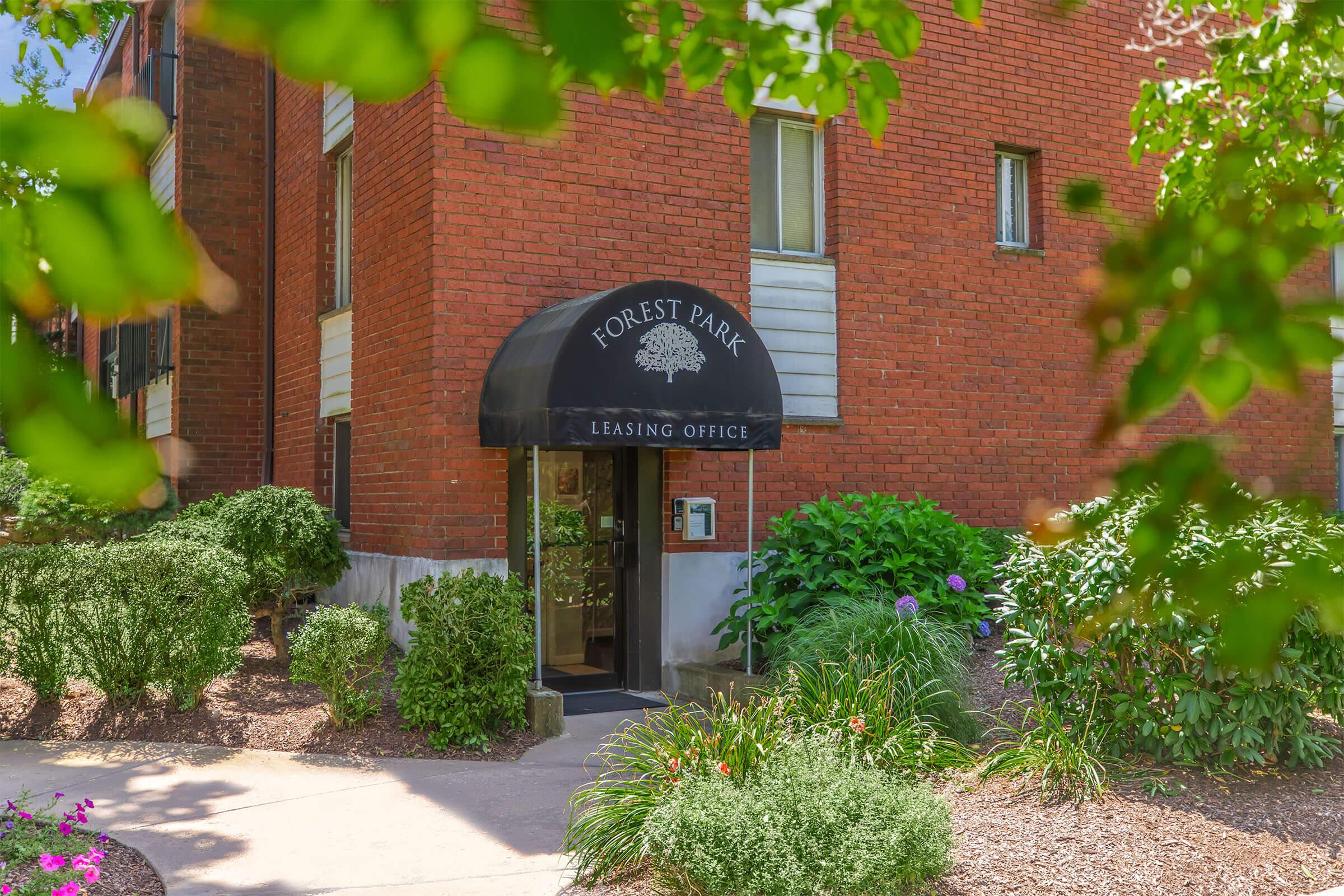 A view of the Forest Park Leasing Office, showcasing a brick building entrance with a black awning. Surrounding greenery includes well-maintained bushes and colorful flowers, providing a welcoming atmosphere. The path leading to the entrance is paved, framed by vibrant plants.