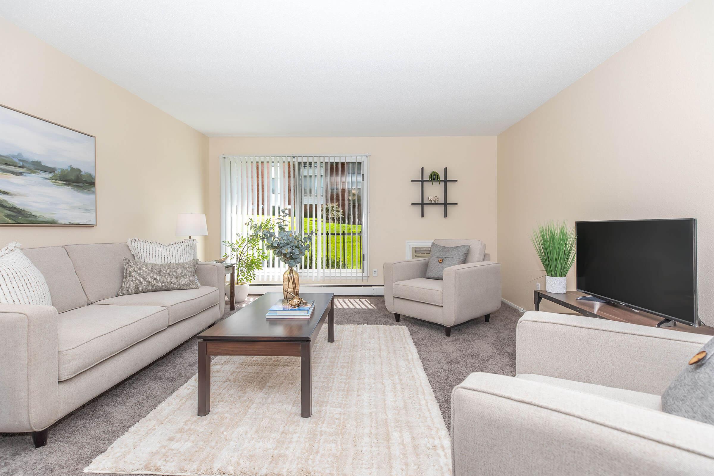 A bright and inviting living room featuring a light-colored sofa and armchairs, a wooden coffee table, and a rug. Large windows allow natural light to flood in, showcasing greenery outside. A small shelf and a television add to the modern decor, creating a cozy and welcoming atmosphere.