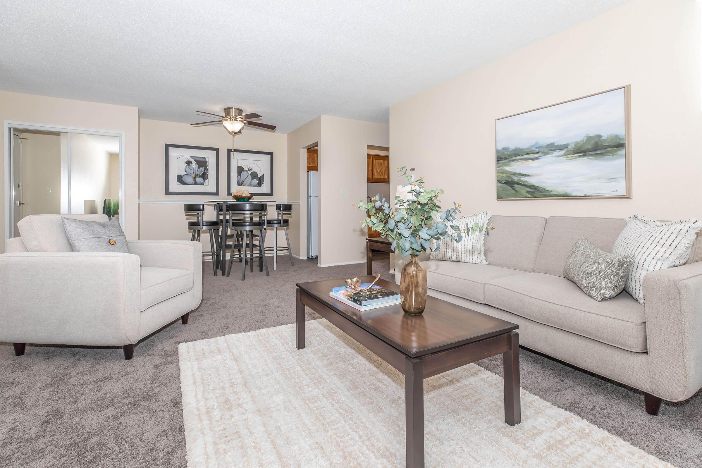 A cozy living room featuring a light-colored sofa and armchair, a wooden coffee table with decorative items, and a soft area rug. Wall art depicts a serene landscape, and there’s a dining area visible in the background. The room has neutral tones, creating a warm and inviting atmosphere.