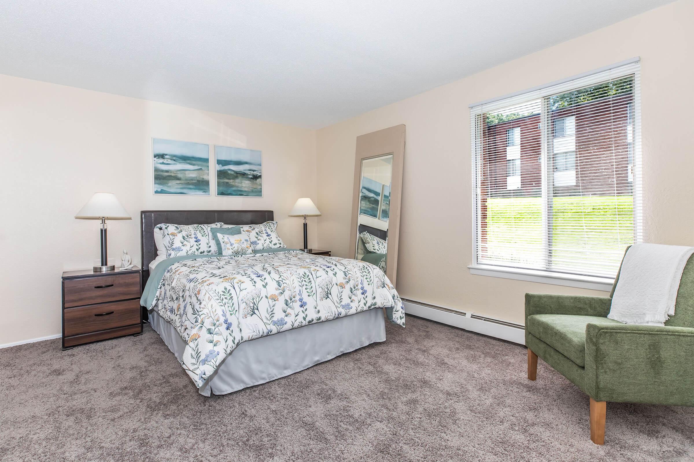 A cozy bedroom featuring a queen-sized bed with a floral-patterned comforter, a bedside table with a lamp, and a large mirror. Natural light streams in through a window with blinds, and a green armchair adds a pop of color to the neutral-toned decor. The carpet is soft and inviting.