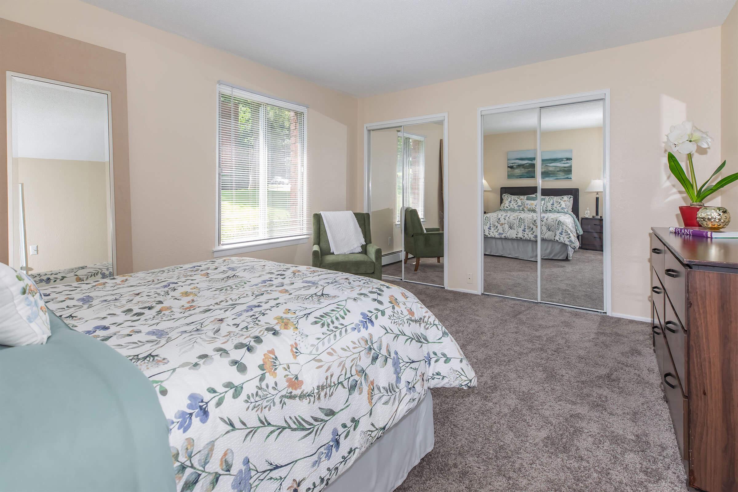 A cozy bedroom featuring a bed with a floral comforter, a large mirror reflecting the room, and light-colored walls. Natural light enters through window blinds. A wooden dresser and two chairs complete the inviting atmosphere. A green plant adds a touch of freshness to the space.