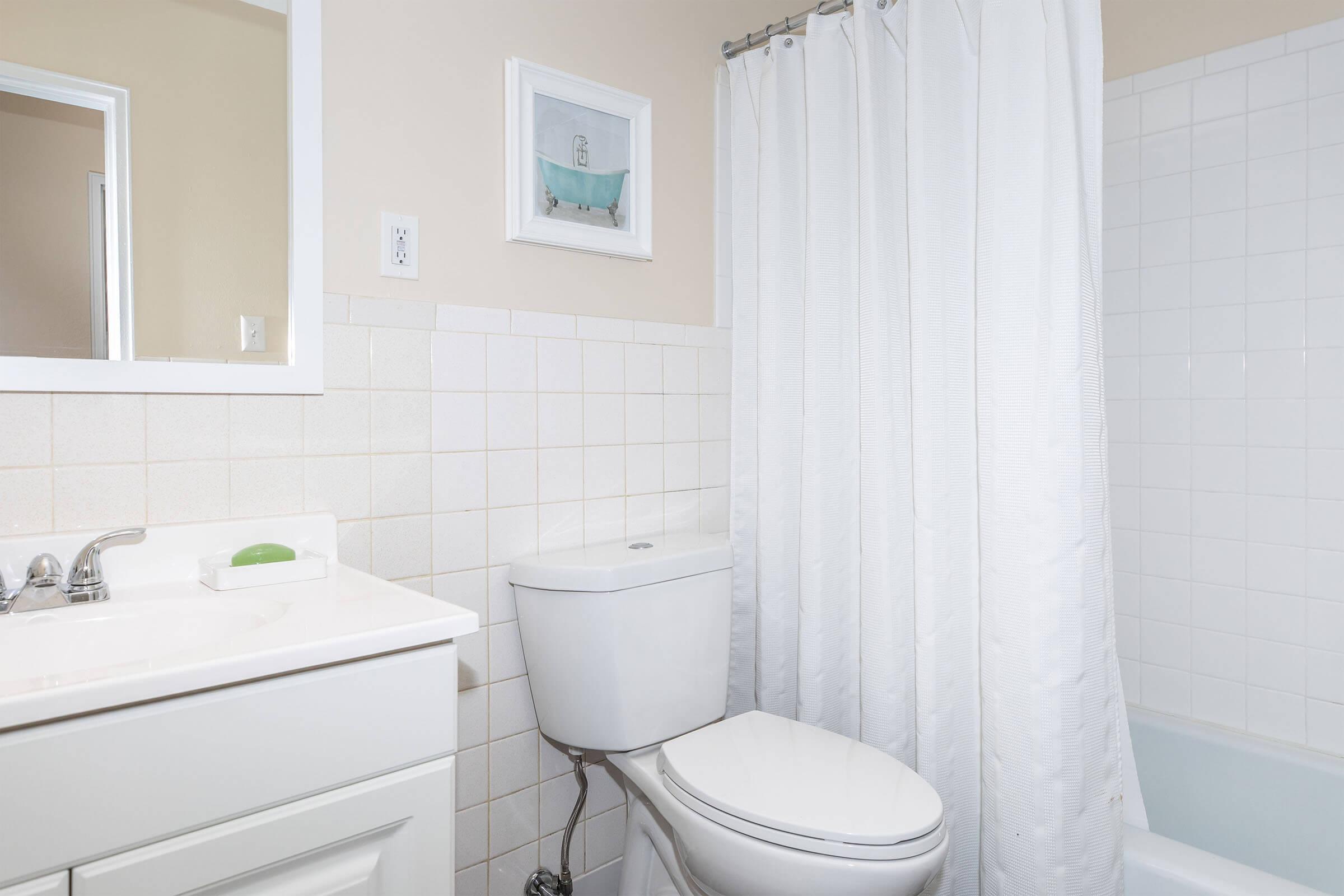 A clean, modern bathroom featuring a white sink with a mirror above, a white toilet, and a bathtub with a shower curtain. The walls are tiled in light colors, and there's a small framed picture on the wall, adding a touch of decor to the space.