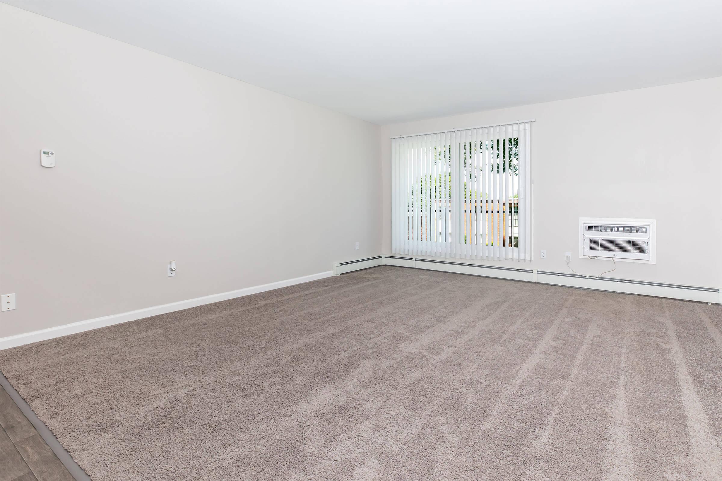 A spacious, empty living room featuring light beige walls and a plush beige carpet. There are vertical blinds covering a large window, allowing natural light to enter. An air conditioning unit is installed beneath the window, and the room has a clean, open feel with minimal furnishings.