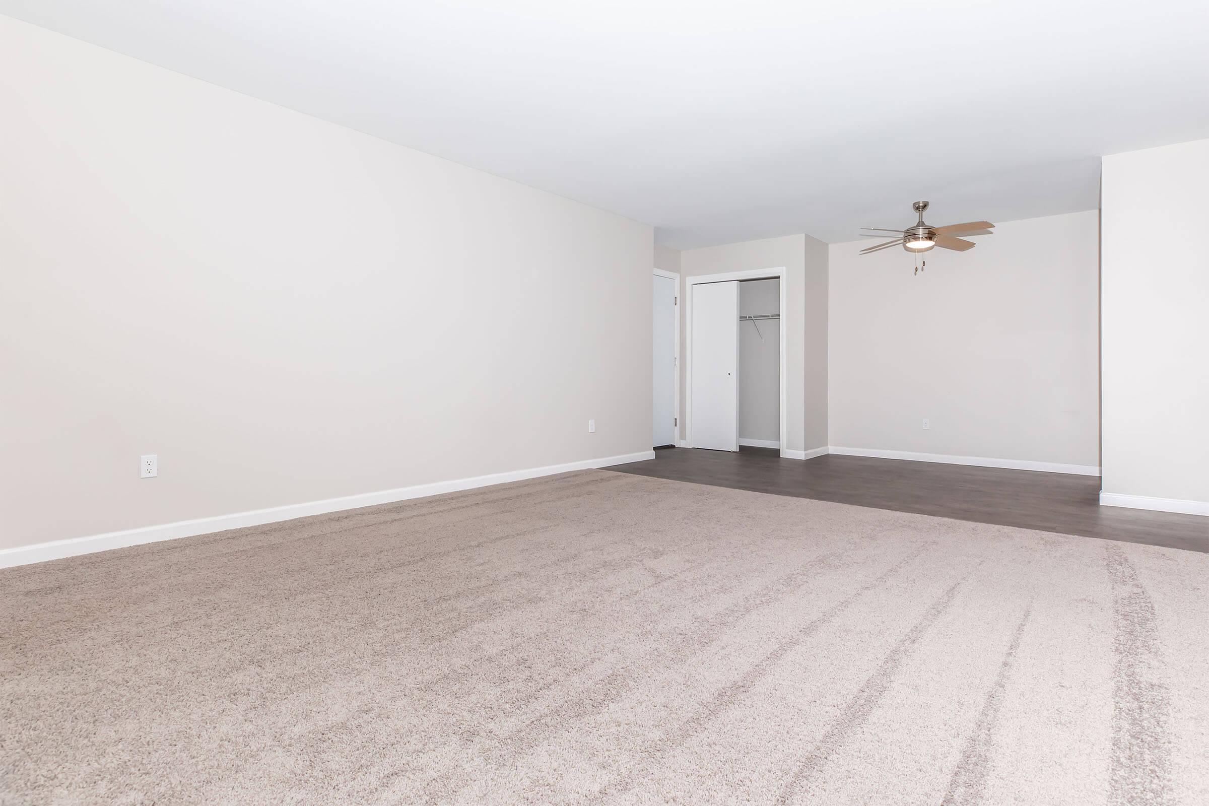 A spacious, empty room with light-colored walls and a beige carpet. There is a ceiling fan in the corner, and a closet door visible on the left side. The floor has a subtle texture, and the overall atmosphere is bright and open.