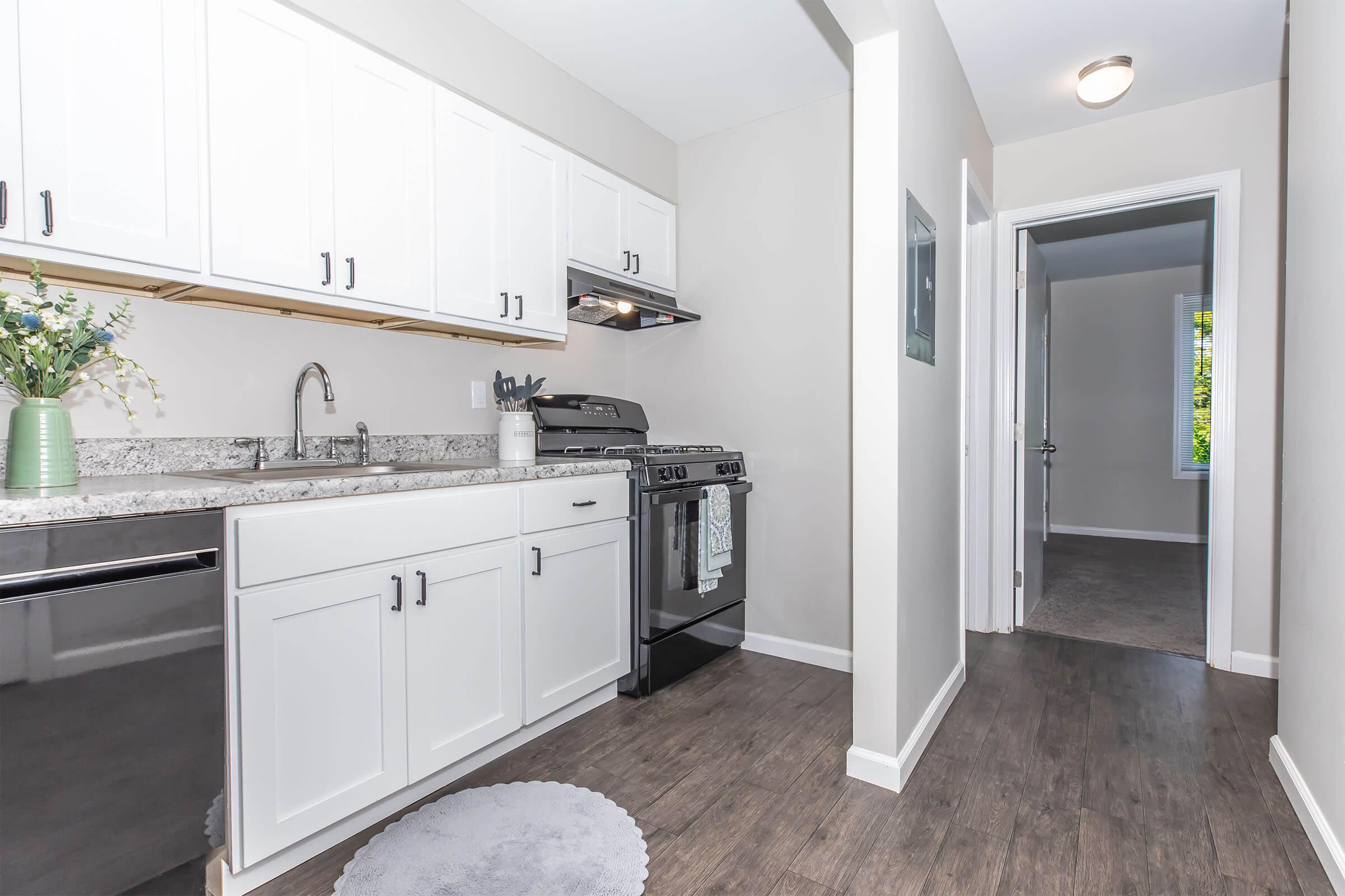 Modern kitchen featuring white cabinetry, granite countertops, and stainless steel appliances. A gas stove is positioned between a sink and a dishwasher. The floor is dark wood, and a round rug is placed in front of the sink. An adjacent hallway leads to a room with a window, adding natural light to the space.