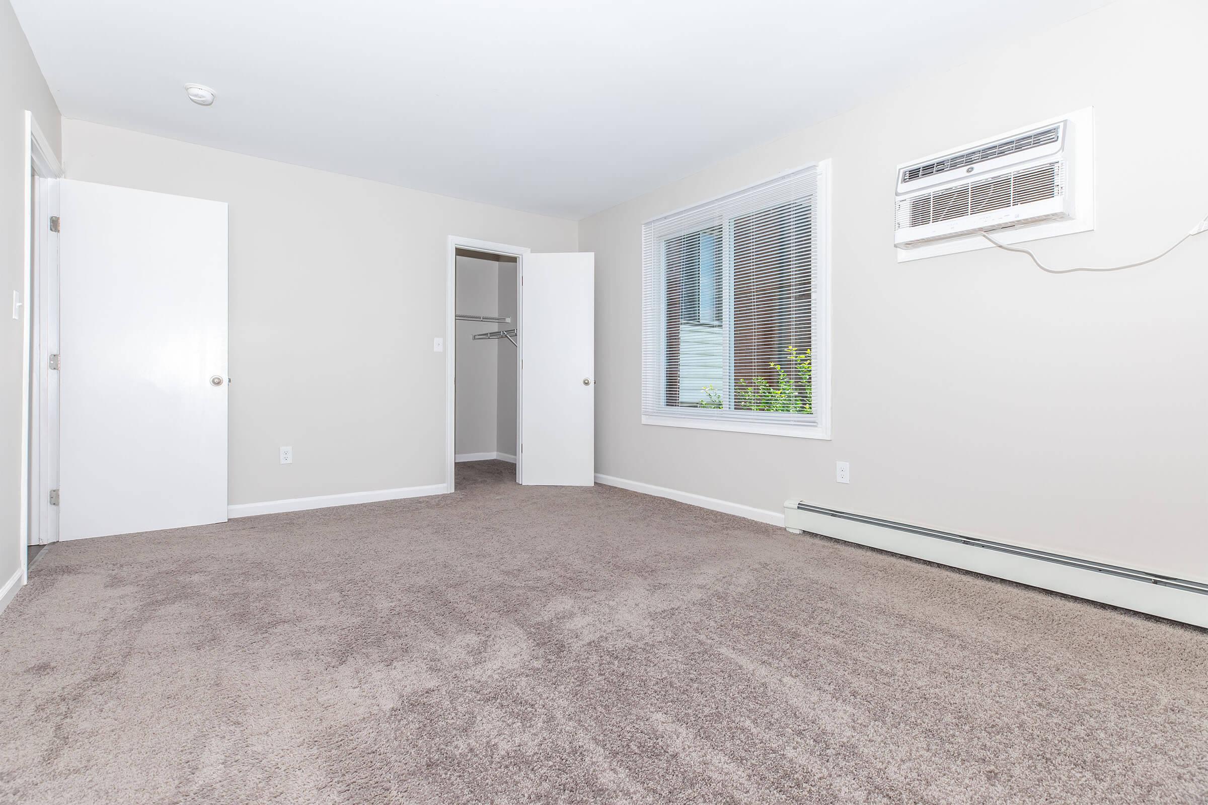 A spacious empty room with light beige walls and plush carpet flooring. It features two closed doors, one leading to a closet, and a window allowing natural light. An air conditioning unit is mounted on the wall. The overall ambiance is bright and clean, suitable for various uses.