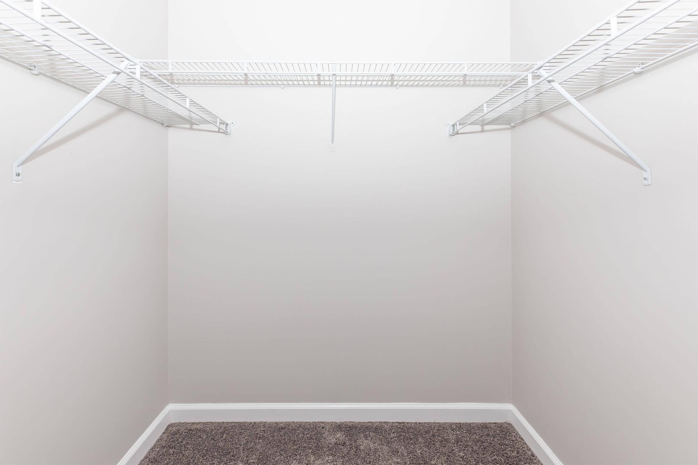 Empty closet interior with light gray walls, featuring a white wire shelving unit along the top. The floor is covered with a dark carpet, and there are no items or additional furniture present in the space. The overall appearance is clean and minimalistic.