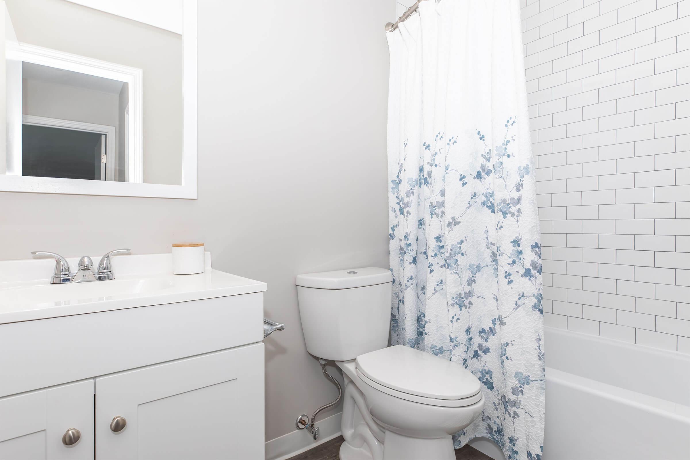 A clean bathroom featuring a white vanity with a sink, a large mirror, a toilet, and a bathtub with a white shower curtain adorned with blue floral patterns. The walls are a soft gray, and the floor has a neutral finish, creating a bright and modern appearance.