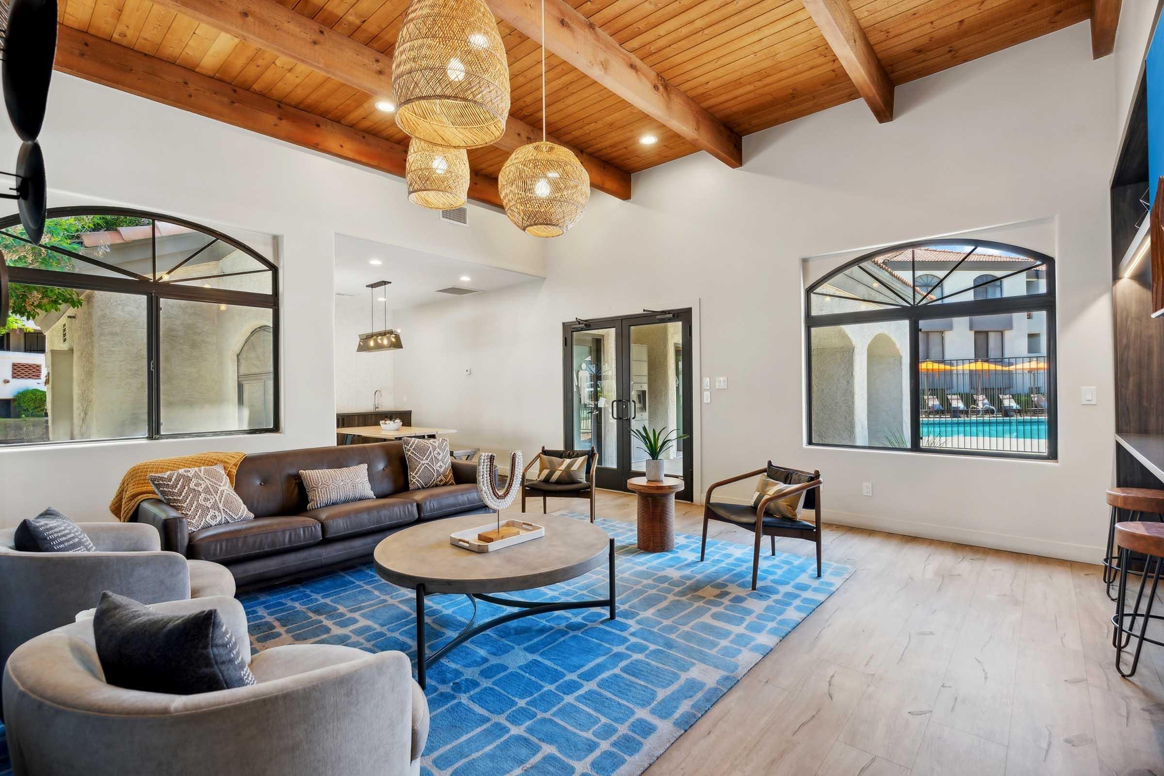 A spacious, modern living room featuring a brown leather sofa, two stylish armchairs, and a round coffee table on a blue patterned rug. The room is adorned with wooden beams on the ceiling and three pendant lights. Large windows allow natural light and a view of a pool area outside.