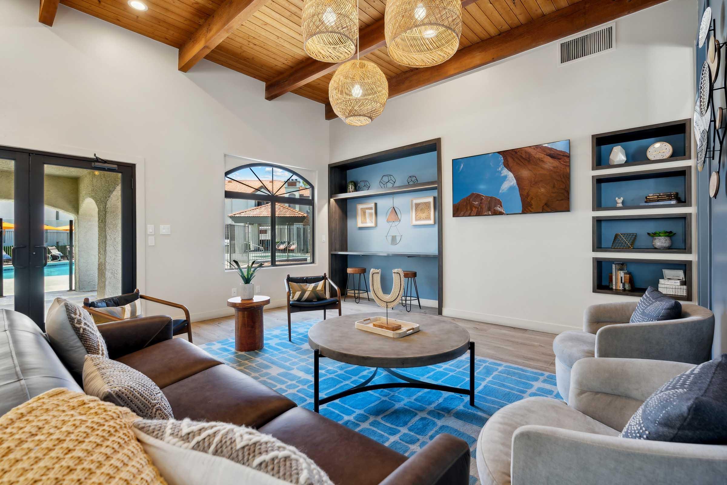A modern living room featuring a high wooden ceiling with three woven pendant lights. There's a comfortable sectional sofa and armchairs surrounding a circular coffee table. A blue area rug covers the floor, and built-in shelves display decorative items. Large windows let in natural light, offering a view of the outdoors.