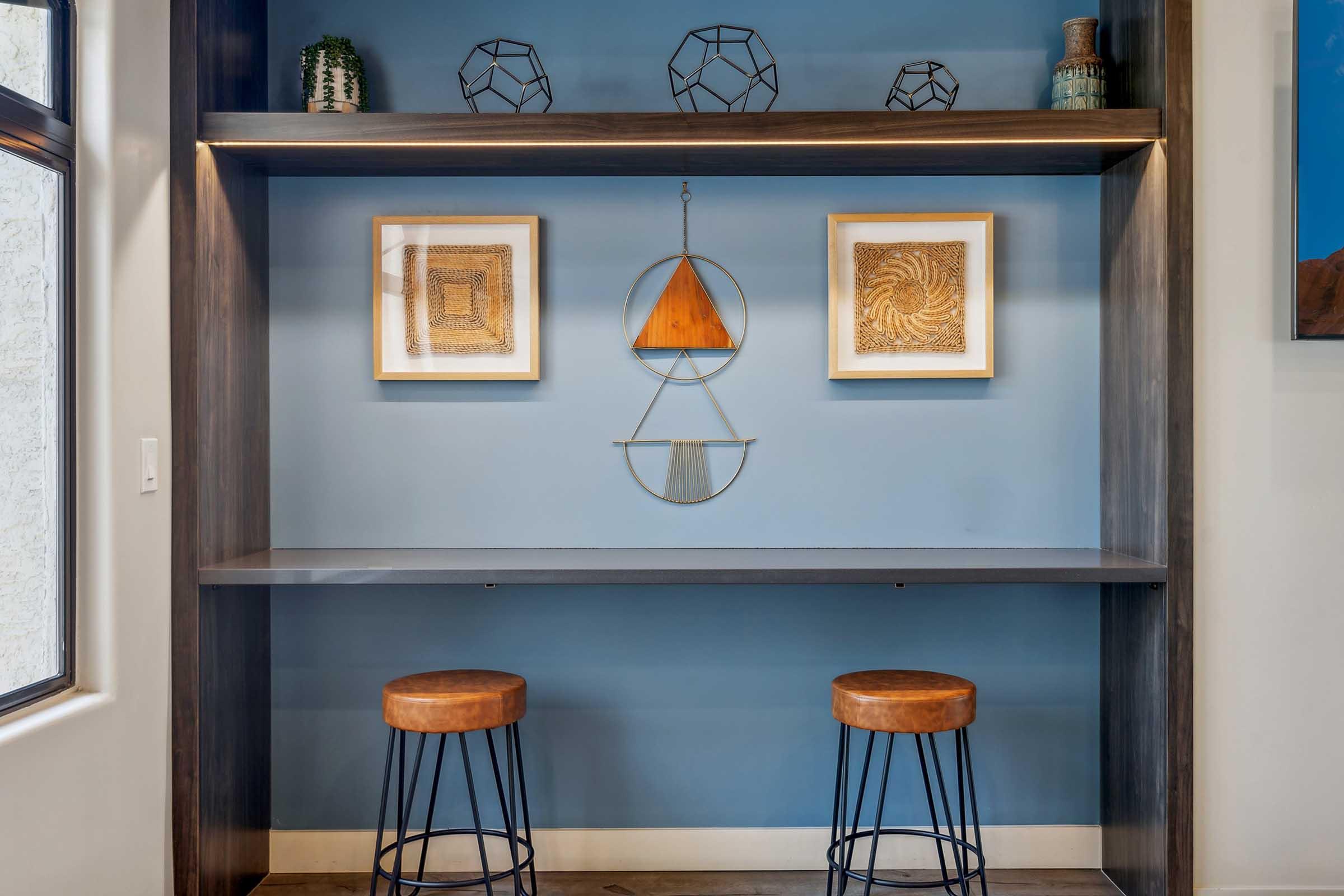 A modern shelving unit against a blue wall featuring two framed artworks, a decorative hanging piece, and three geometric sculptures. Below, two stylish bar stools with wooden seats. The overall aesthetic is contemporary and organized.