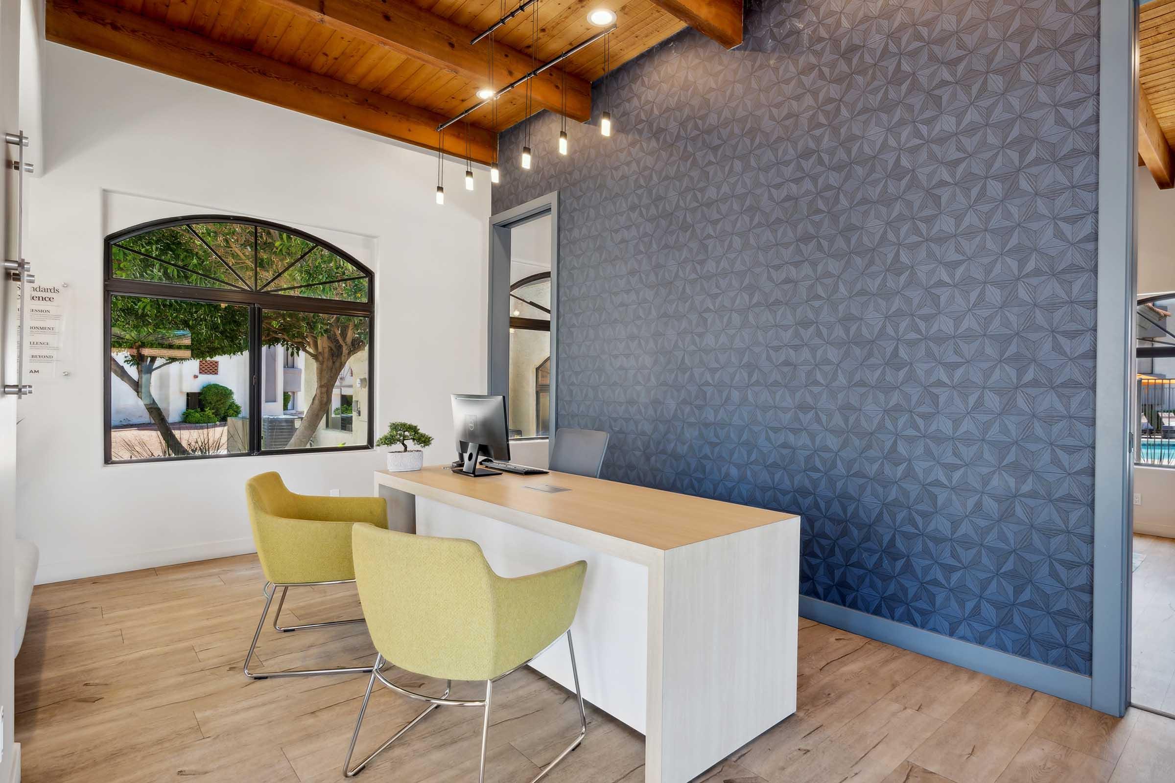 A modern office space featuring a light wooden desk with a computer and two green chairs. The walls are painted light colors, with a textured blue accent wall. Large windows allow natural light in, and a view of greenery is visible outside. The ceiling has wooden beams and pendant lighting.