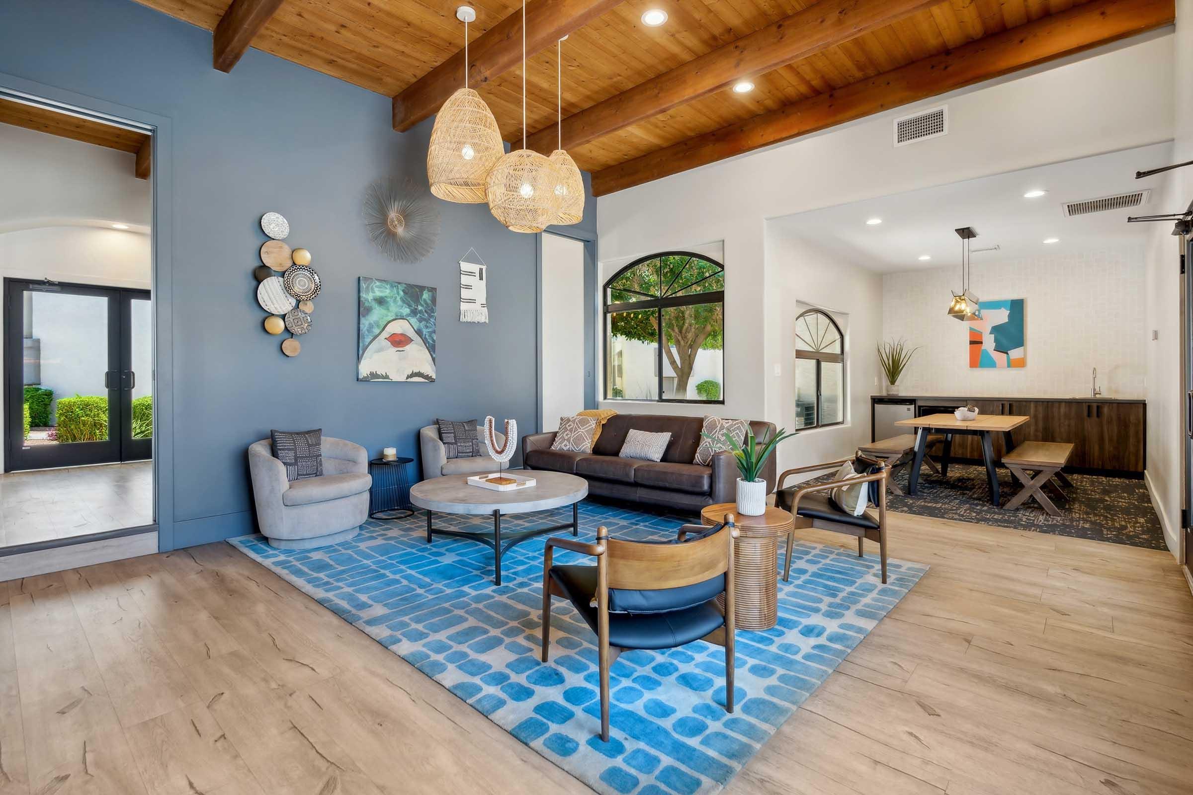 A modern living room featuring an open layout with wooden beams, a cozy seating area including a gray sofa and chairs, a round coffee table, and decorative wall art. Natural light streams in through large windows, creating a bright atmosphere. The flooring is light wood, complementing the vibrant blue area rug.
