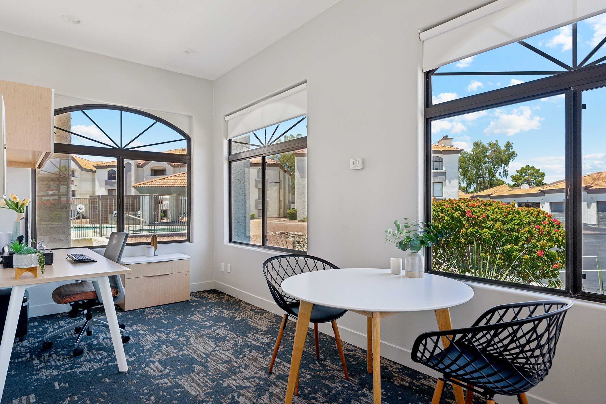 A bright, modern workspace featuring a small round table and chairs near large windows. The windows offer a view of a landscaped outdoor area with greenery and a pool. A desk with a chair is positioned against one wall, creating a cozy, inviting atmosphere.