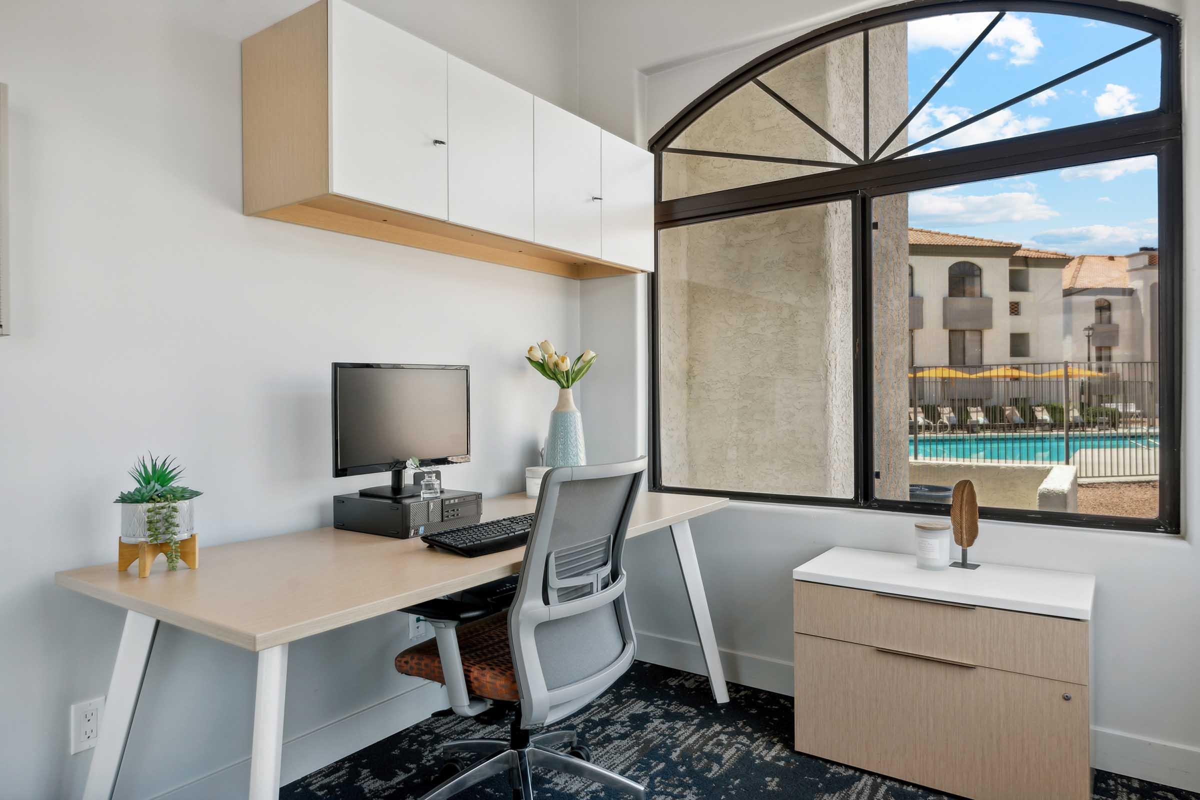 A modern workspace featuring a desk with a computer, ergonomic chair, and decorative plant. The room has large windows providing a view of a pool area outside, with a light, airy atmosphere enhanced by natural light. Storage cabinets are mounted above the desk, and a small side table is nearby.