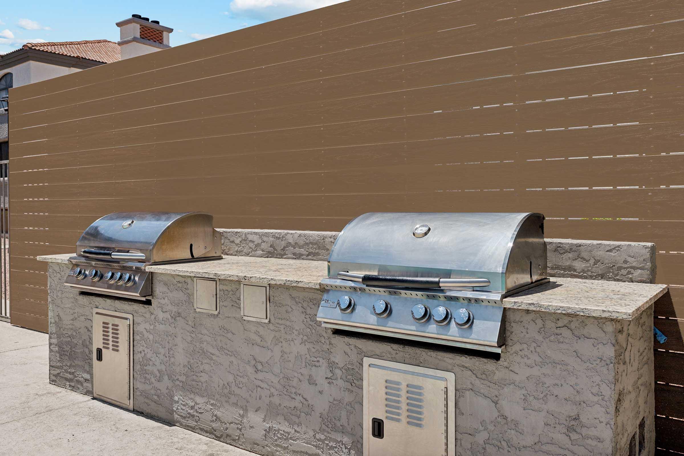 Two stainless steel outdoor grills on a stone countertop, placed against a wooden privacy fence. The grills have shiny domed lids and control knobs, with storage compartments below. The area is well-lit and spacious, suitable for outdoor cooking and entertaining.