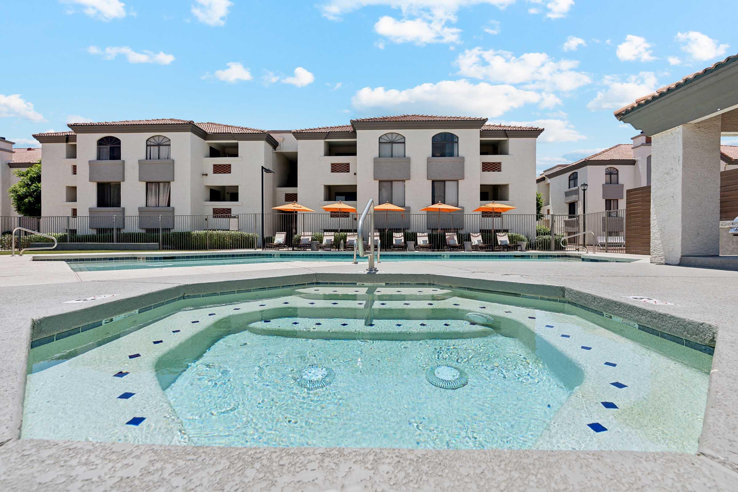 A bright outdoor pool area featuring a hot tub in the foreground, surrounded by lounge chairs with orange umbrellas. In the background, a multi-story residential building is visible under a clear blue sky with a few clouds. The scene conveys a relaxing atmosphere ideal for leisure and recreation.