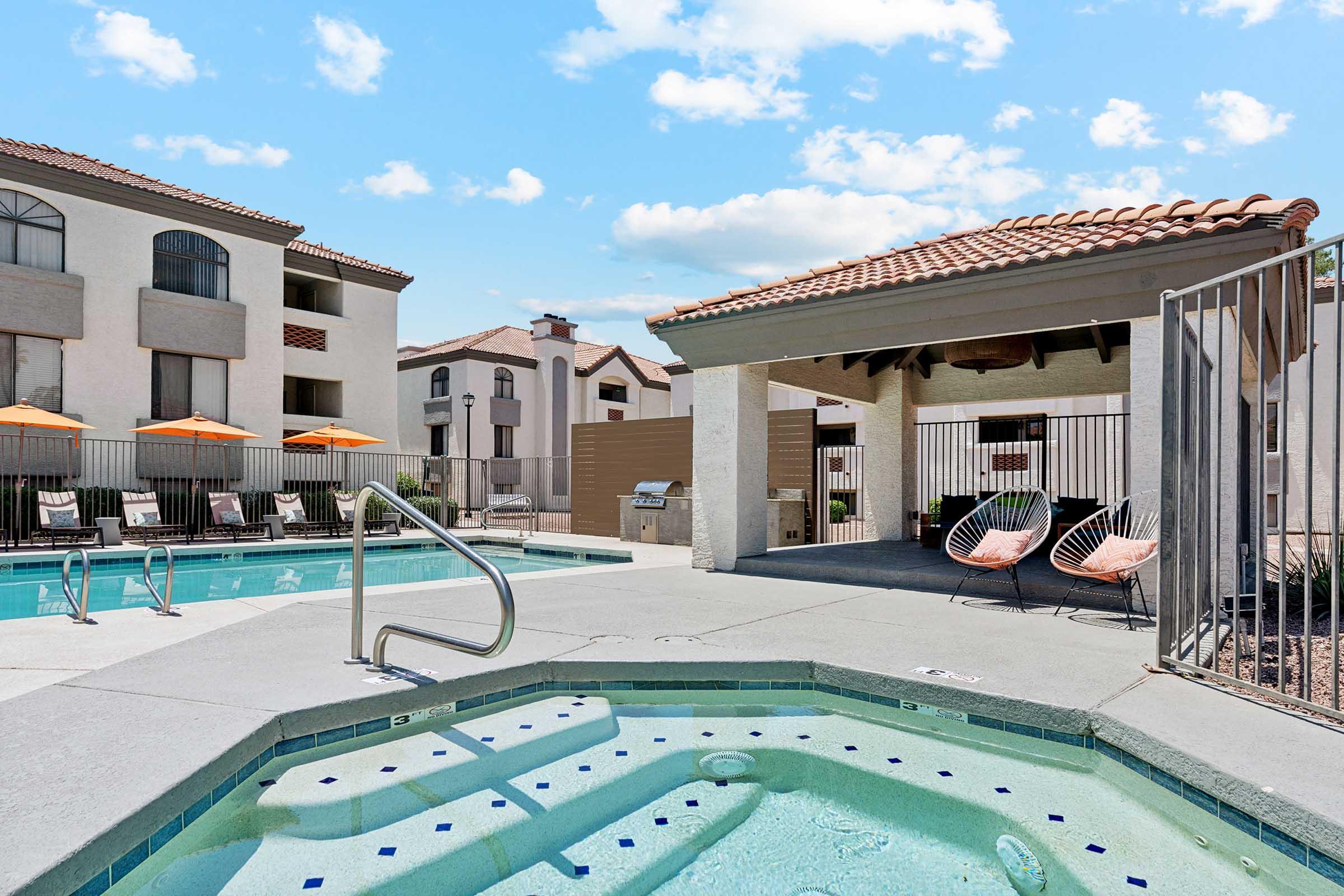 Swimming pool area featuring a hot tub, lounge chairs, and orange umbrellas. A shaded seating space with modern chairs is visible in the background. The sky is bright with fluffy clouds, and the surrounding buildings have a Mediterranean architectural style.