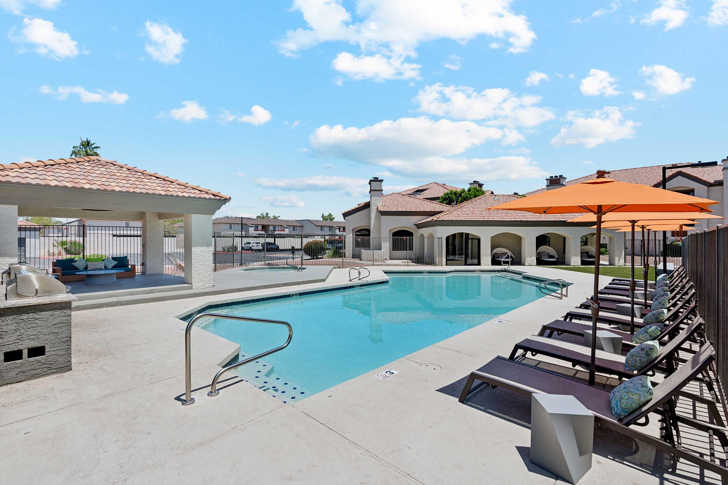 A bright outdoor swimming pool area featuring a clear blue pool surrounded by lounge chairs and orange umbrellas. In the background, there's a residential building with a tiled roof, and the scene is set against a bright sky with fluffy clouds.
