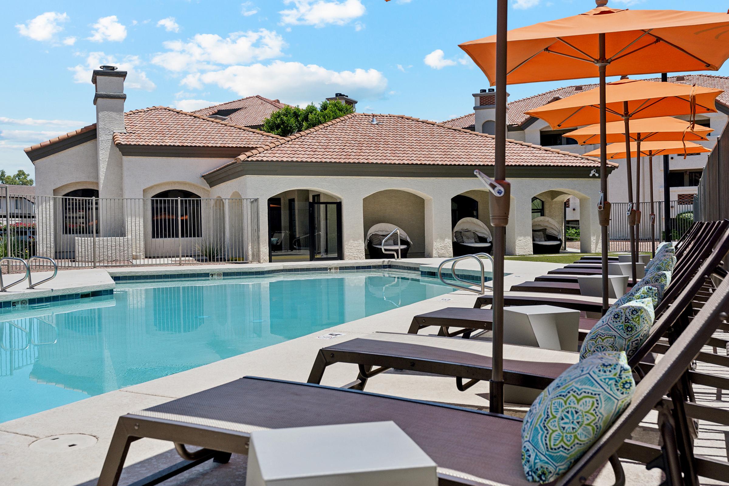 A sunny day by a pool area featuring lounge chairs with decorative pillows, orange umbrellas, and a surrounding fence. In the background, there is a stylish building with a red-tiled roof. The scene is bright and inviting, perfect for relaxation.
