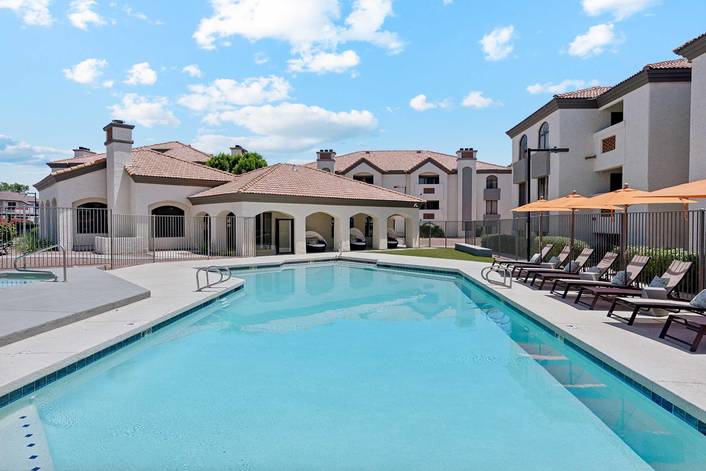 A serene pool area surrounded by lounge chairs, with an adjacent building featuring a Spanish-style architecture. The pool has clear blue water, and orange umbrellas provide shade. The sky is partly cloudy, creating a bright and inviting atmosphere.