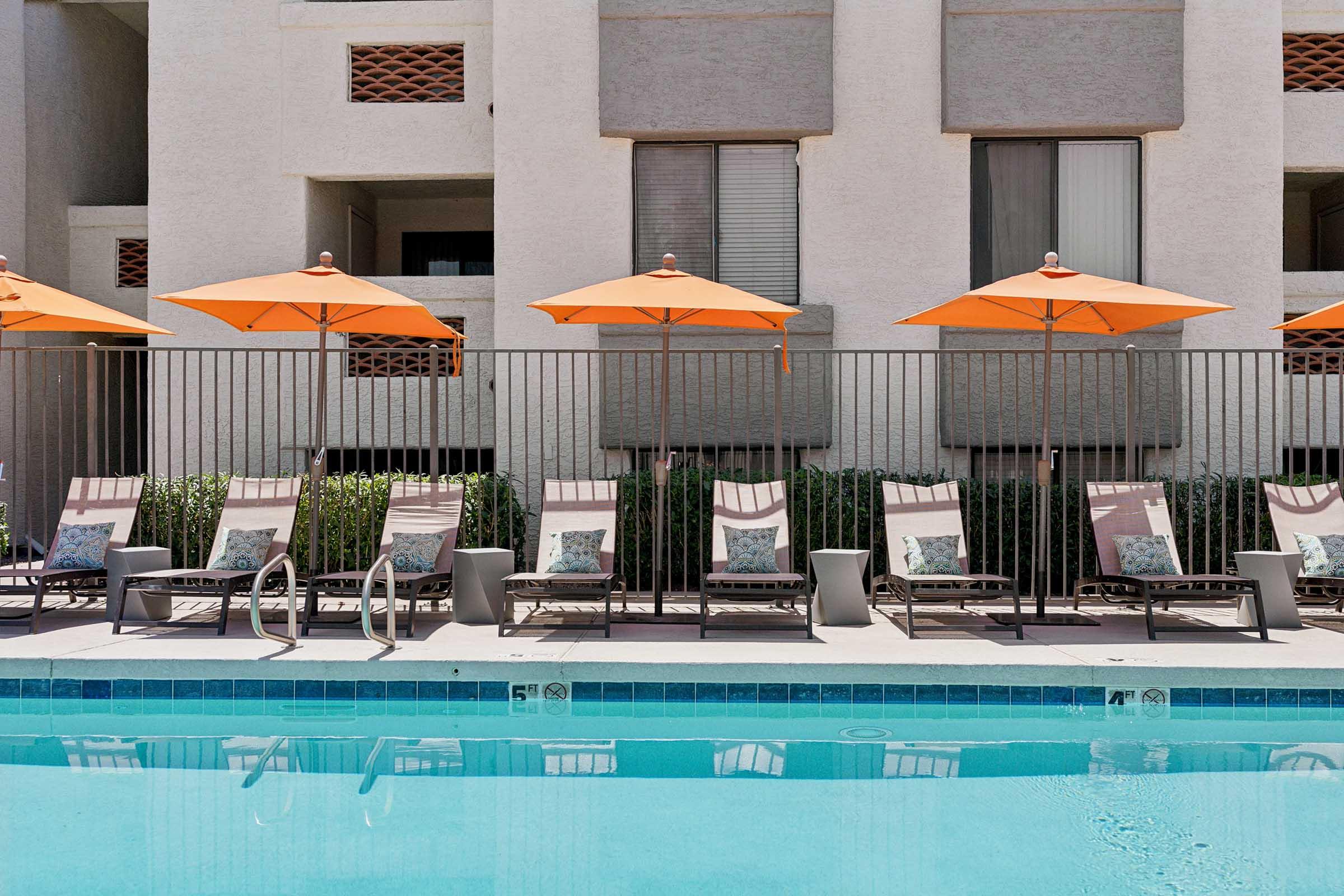 A swimming pool with clear blue water, surrounded by lounge chairs under orange umbrellas. The area features a fence separating the pool from a building with large windows and a neutral-colored exterior. Lush greenery is visible along the poolside.