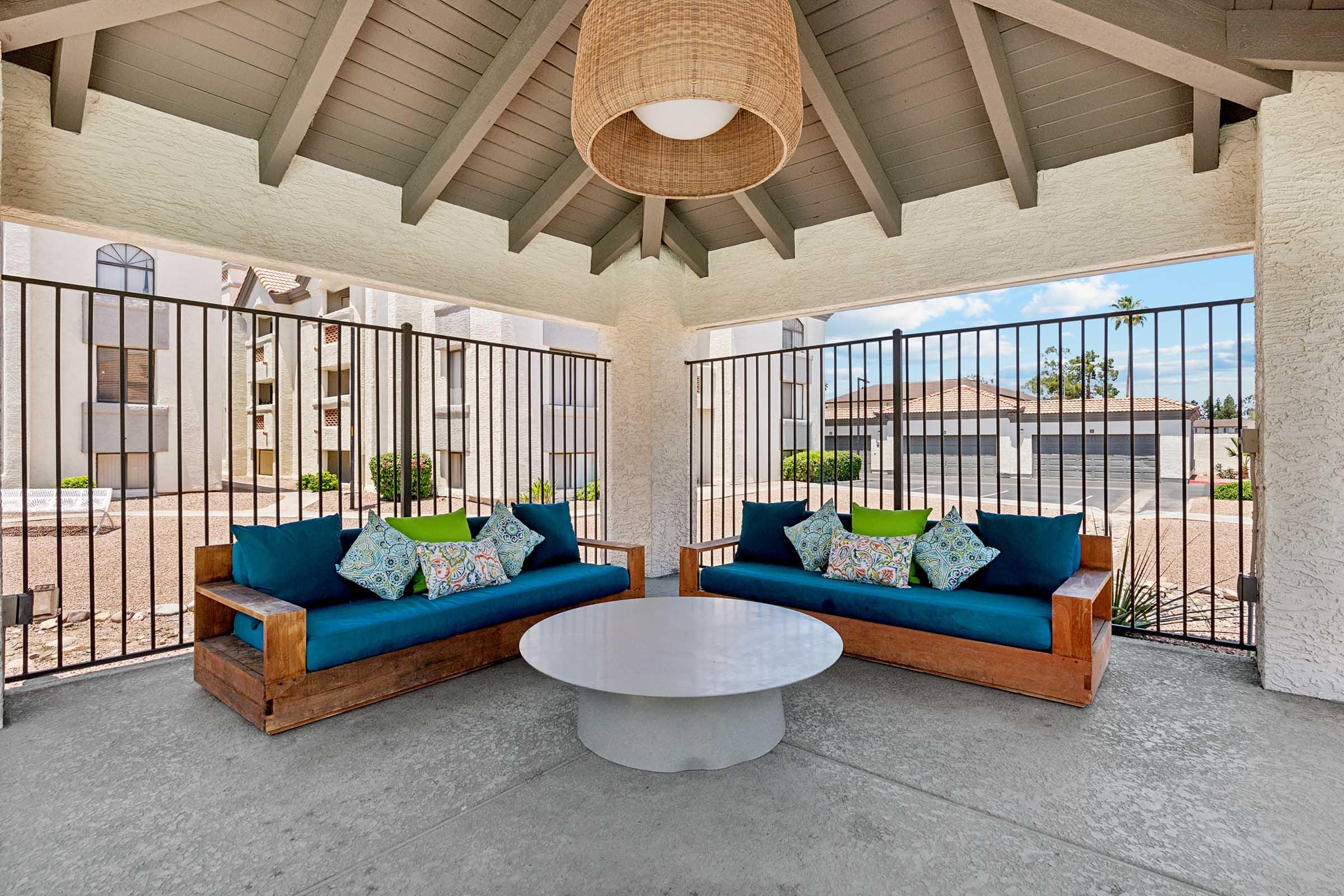 A cozy outdoor seating area featuring two wooden couches with blue cushions and colorful throw pillows, alongside a round white coffee table. The space is sheltered by a high ceiling with wooden beams and a large light fixture, surrounded by decorative metal fencing, with greenery and buildings in the background.