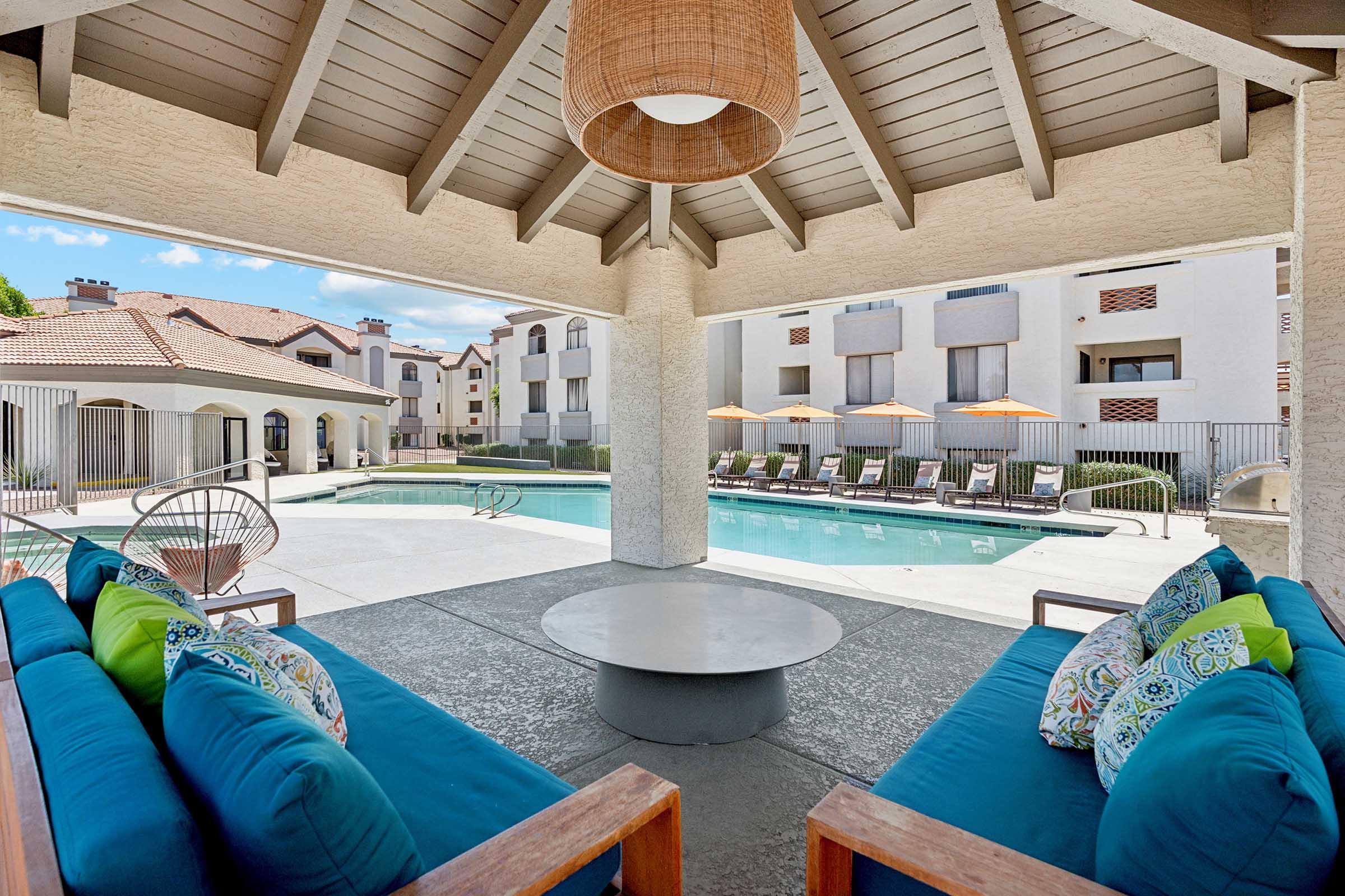 Covered lounge area with comfortable, vibrant blue sofas adorned with patterned pillows. A round coffee table sits in the center, and a swimming pool is visible in the background, surrounded by sun loungers and orange umbrellas. The setting features a relaxed atmosphere with modern architecture.
