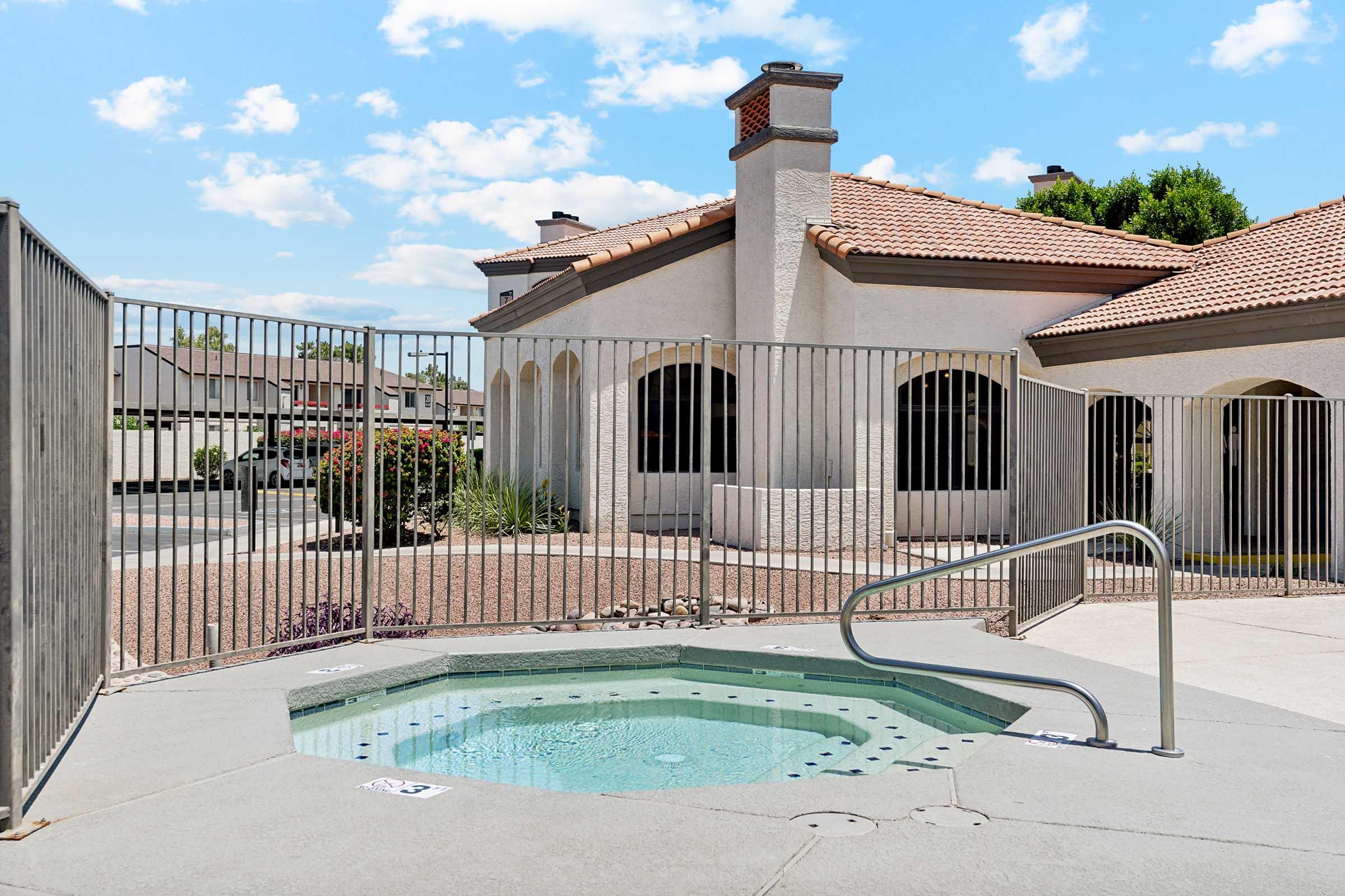A small outdoor hot tub surrounded by a metal fence, situated next to a single-story house with a tiled roof. The sky is bright with scattered clouds, and there are some shrubs and trees in the background. The area is well-maintained with a patio surface.