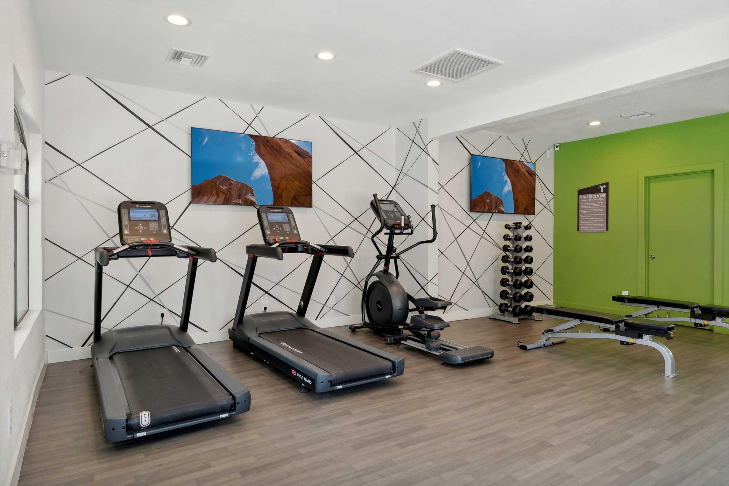 A fitness room featuring two treadmills, an elliptical machine, and a weight bench with free weights. The walls are adorned with modern geometric patterns and landscape artwork. There is a vibrant green wall, enhancing the bright and inviting atmosphere of the space.