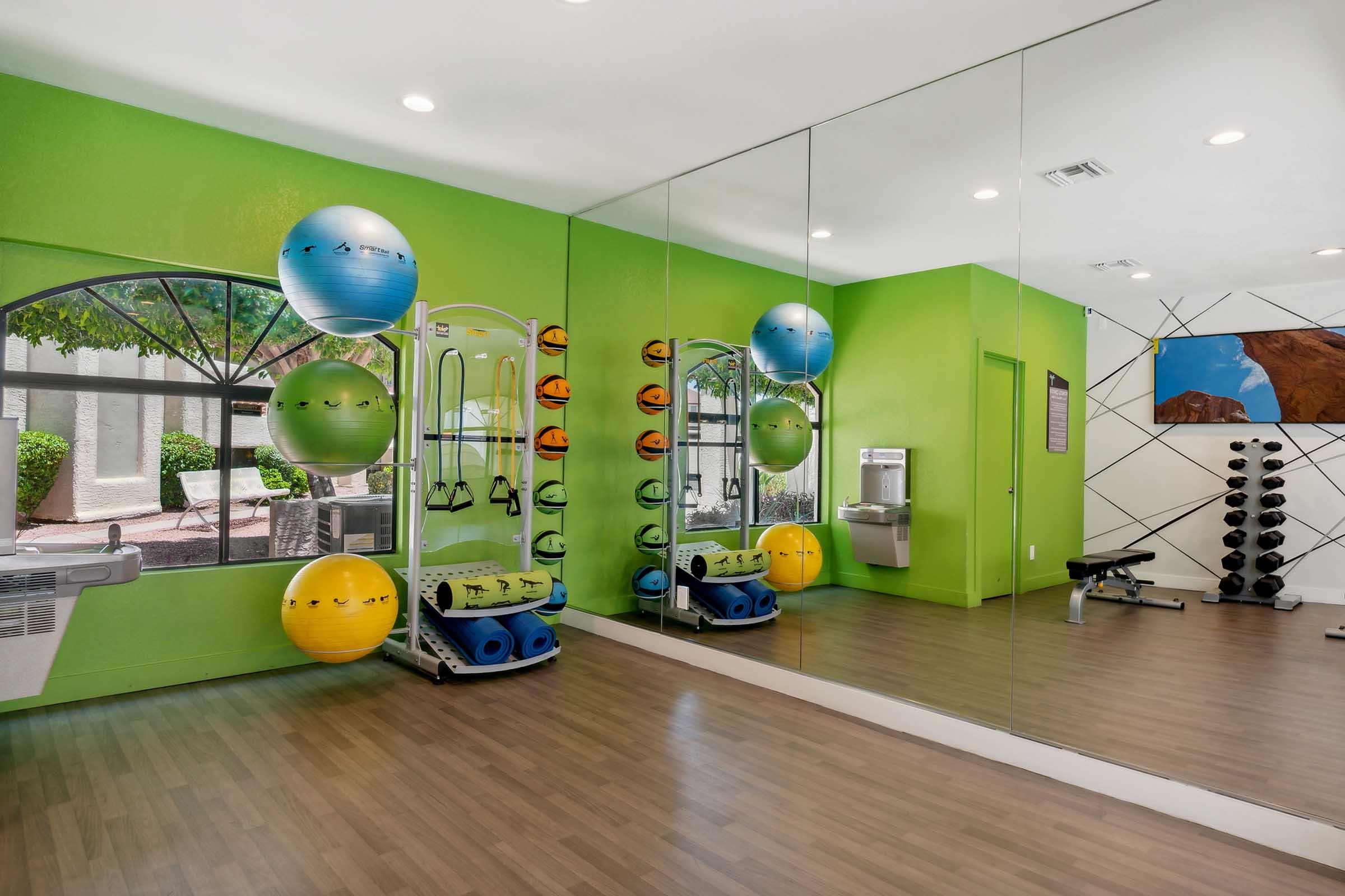 Brightly colored gym interior featuring a green wall, large mirrors, and fitness equipment. There are exercise balls arranged on a wall, a water fountain, and a weight bench with dumbbells. The space is well-lit and organized for workouts.