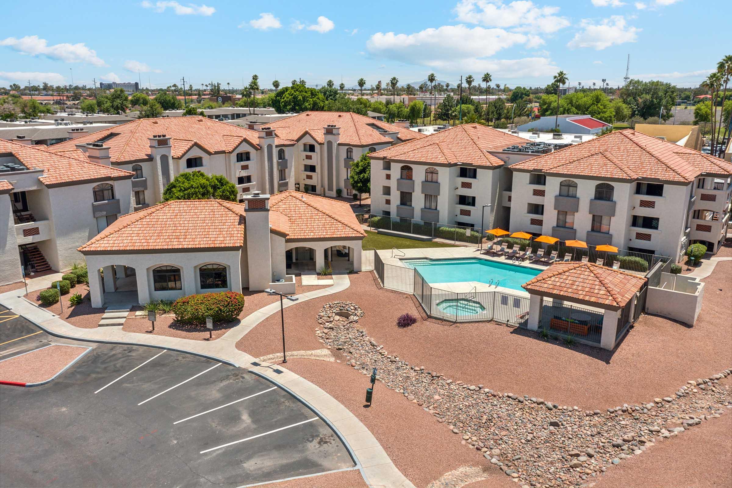 Aerial view of a residential complex featuring multiple buildings with red-tiled roofs, a swimming pool surrounded by lounge chairs, and landscaped areas with rocky paths. The parking lot is visible in the foreground, along with palm trees and blue skies in the background.