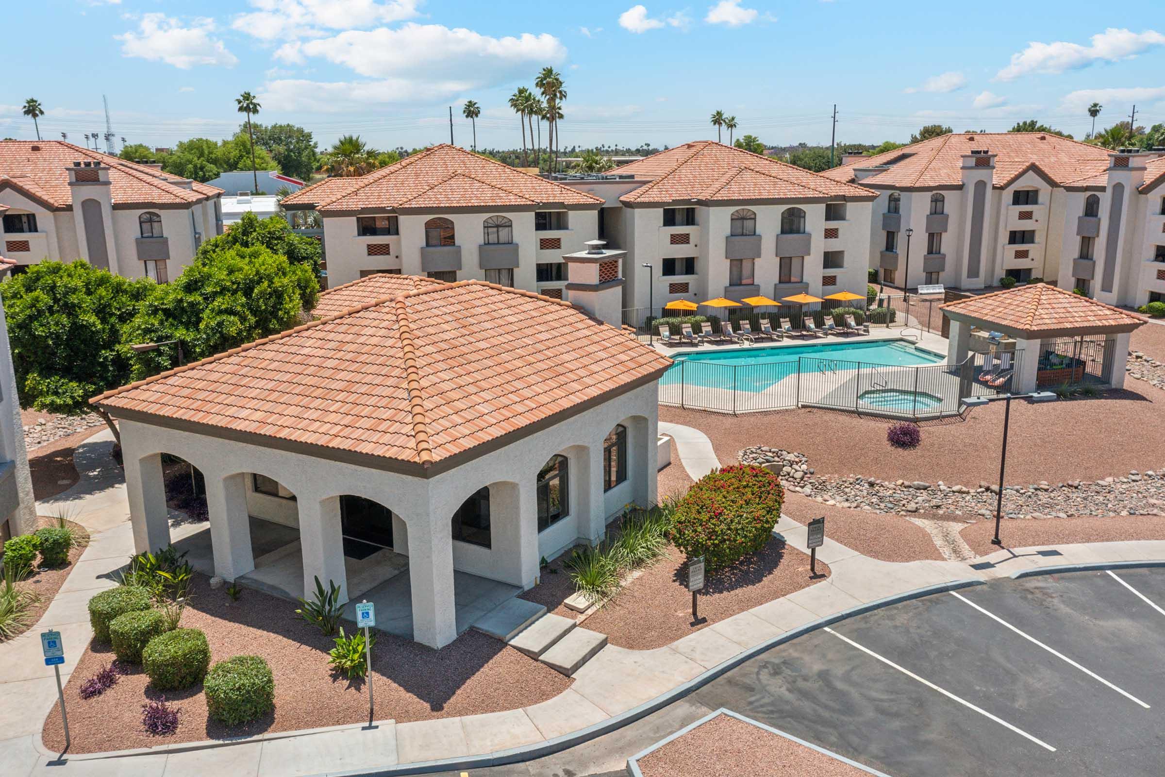 A view of a hotel complex featuring a swimming pool surrounded by lounge chairs. The building has a tan exterior with terracotta roof tiles and lush landscaping including palm trees and shrubs. The area appears sunny and well-maintained with a parking lot in the foreground.
