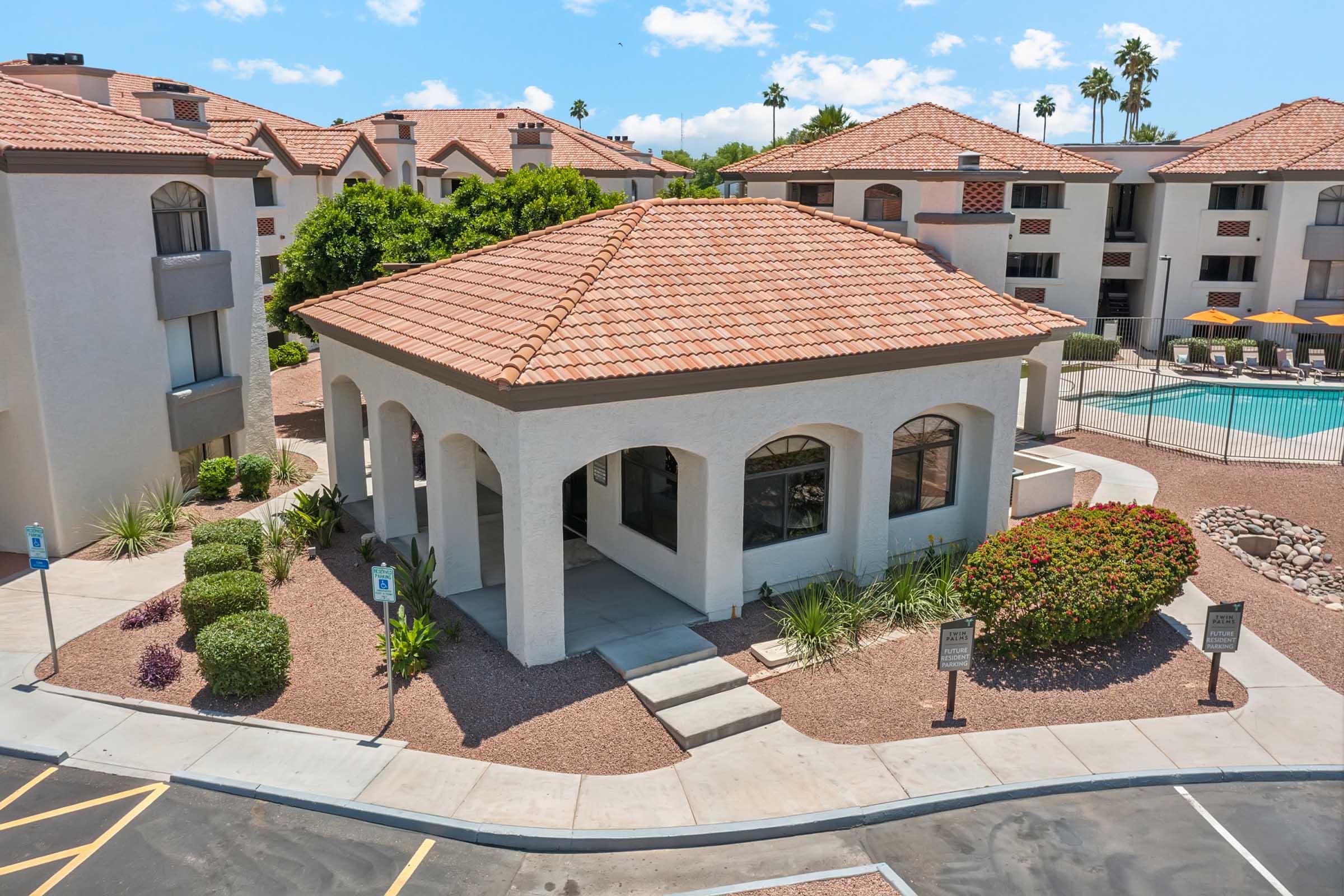 A modern, single-story building with a terracotta tiled roof, surrounded by landscaped gardens and shrubs. In the background, multiple apartment buildings are visible, along with a swimming pool and palm trees under a clear blue sky. The scene is well-lit and captures a sunny day.
