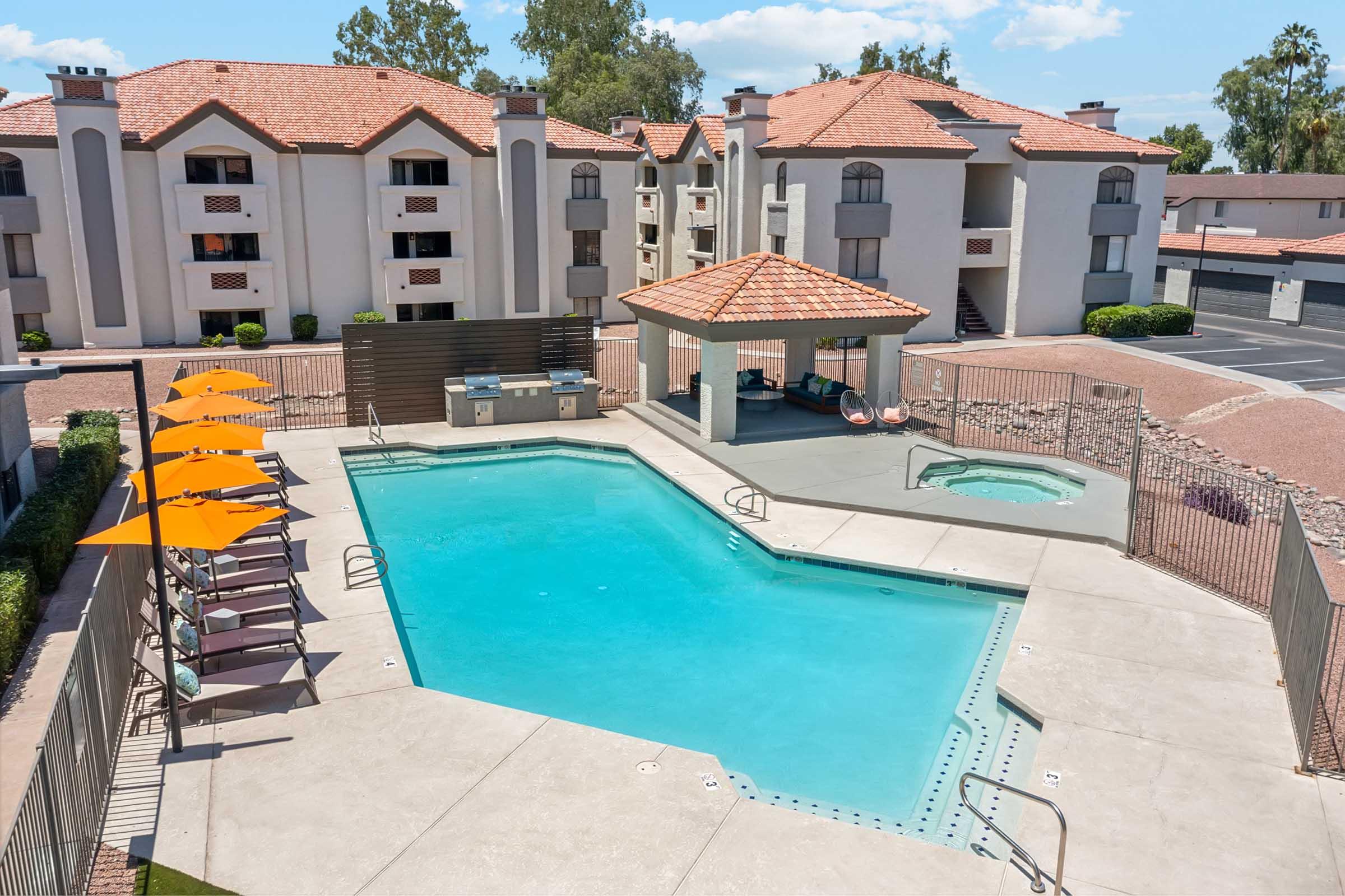 Aerial view of a swimming pool surrounded by lounge chairs with orange umbrellas in a landscaped area. In the background, there are multi-story apartment buildings with red-tiled roofs. A hot tub is visible next to the pool, and there is a shaded seating area nearby. Clear skies and green trees complete the scene.