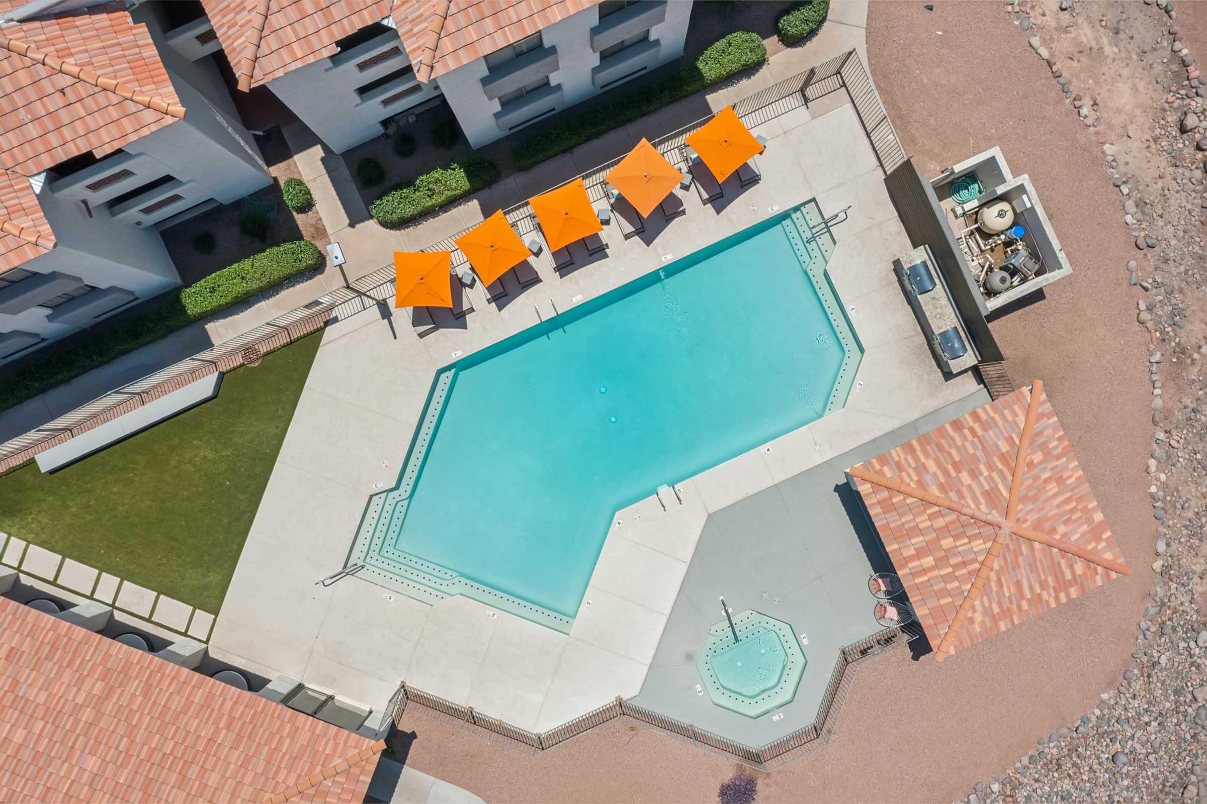 Aerial view of a swimming pool with an octagonal shape, surrounded by orange umbrellas and lounging areas. Nearby, there are landscaped areas and outdoor features, including a grill station and a small fountain, against a backdrop of residential buildings with tiled roofs.