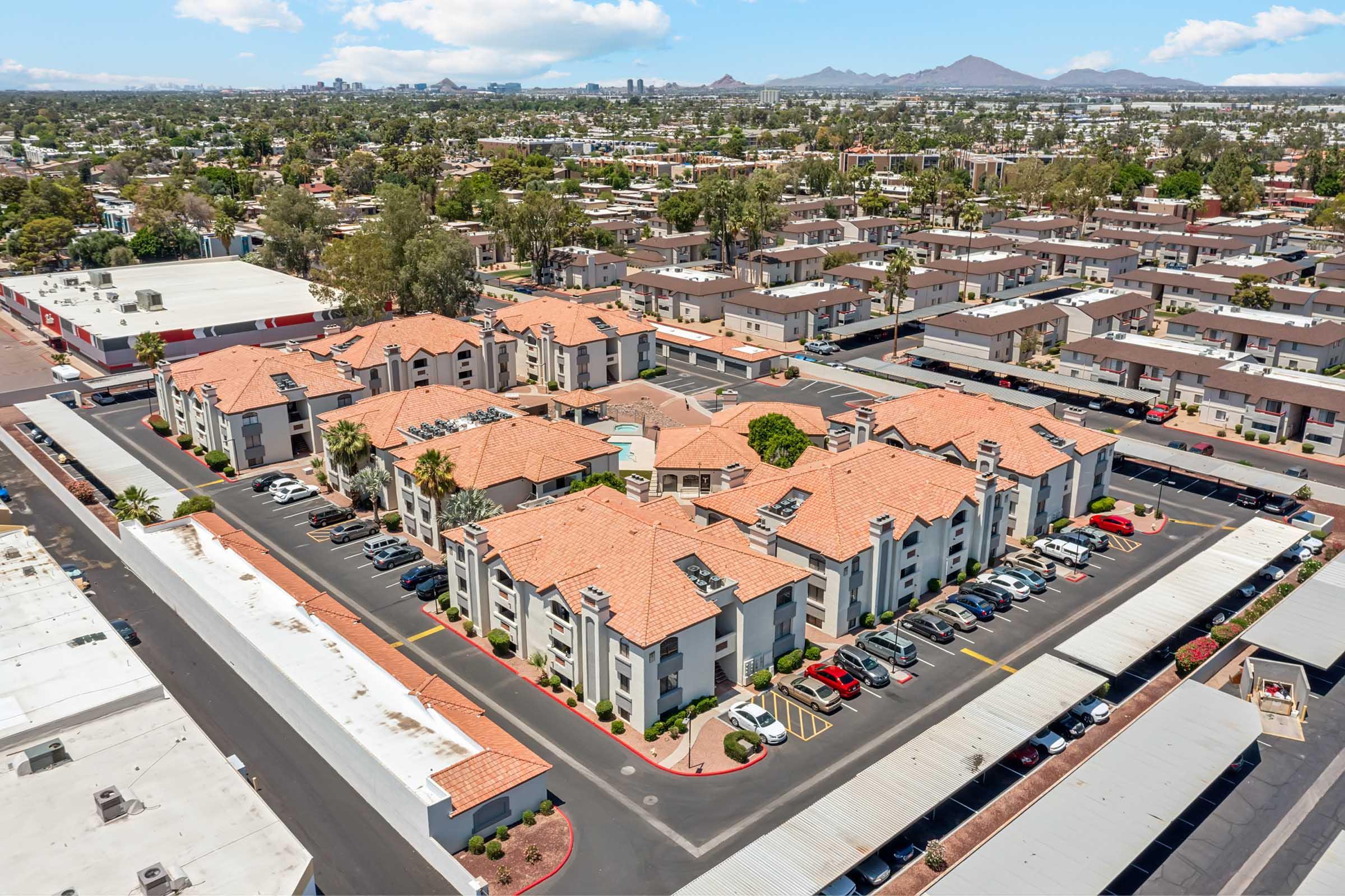 Aerial view of a residential apartment complex featuring multiple buildings with red tiled roofs surrounded by parking areas. Nearby structures and a cityscape can be seen in the background under a blue sky with scattered clouds.