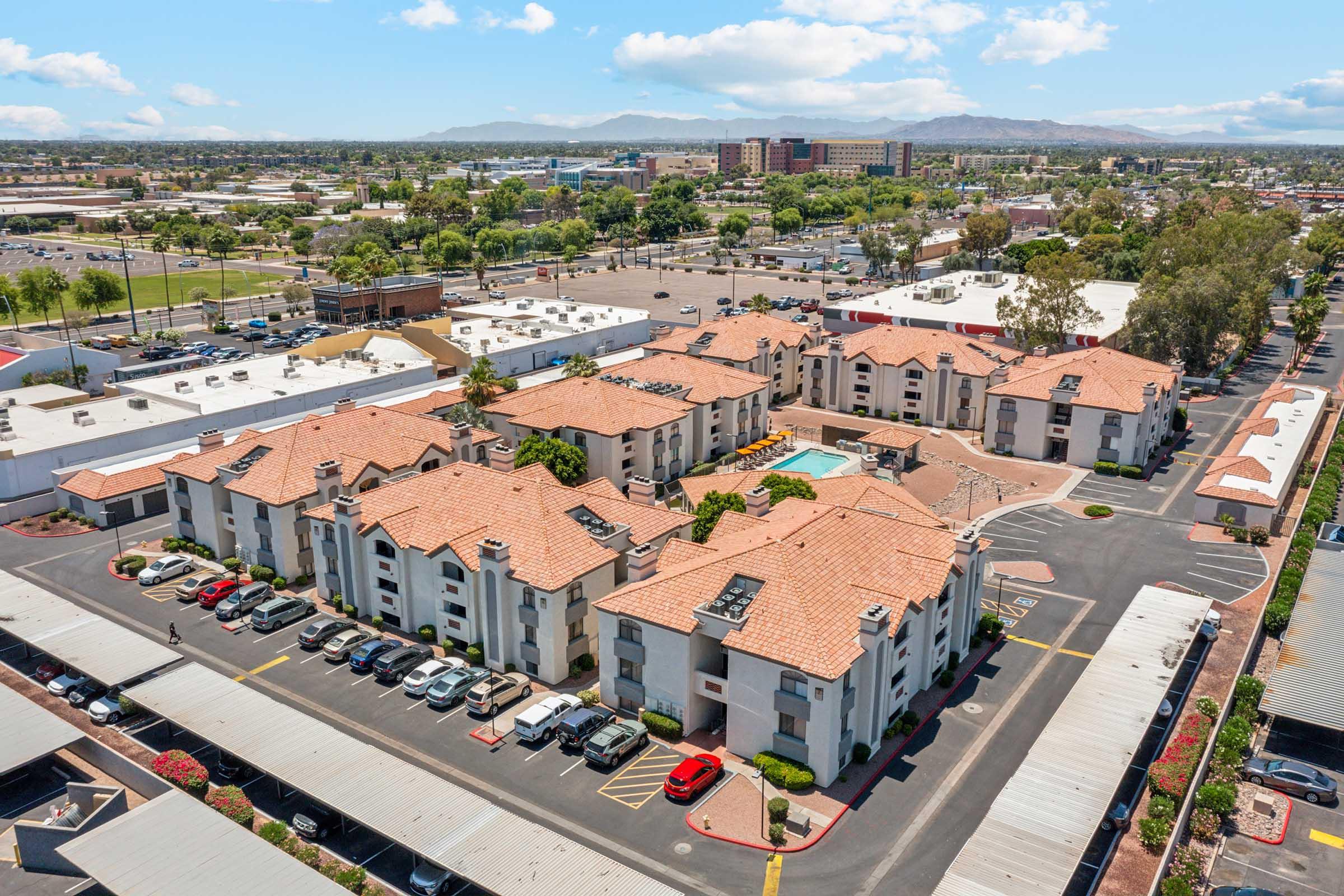 Aerial view of a residential complex featuring multiple buildings with red-tiled roofs, surrounded by parked cars. In the foreground, there is a landscaped area with a swimming pool. The background shows a commercial area and distant mountains under a clear blue sky.