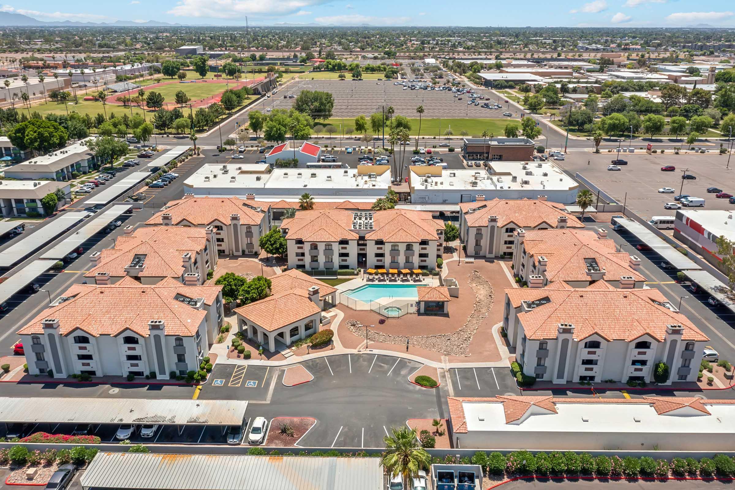 Aerial view of a residential complex featuring multiple buildings with red-tiled roofs, a central swimming pool, and landscaped outdoor areas. Surrounding the complex are parking spaces and nearby commercial buildings, with a backdrop of greenery and distant hills under a clear blue sky.
