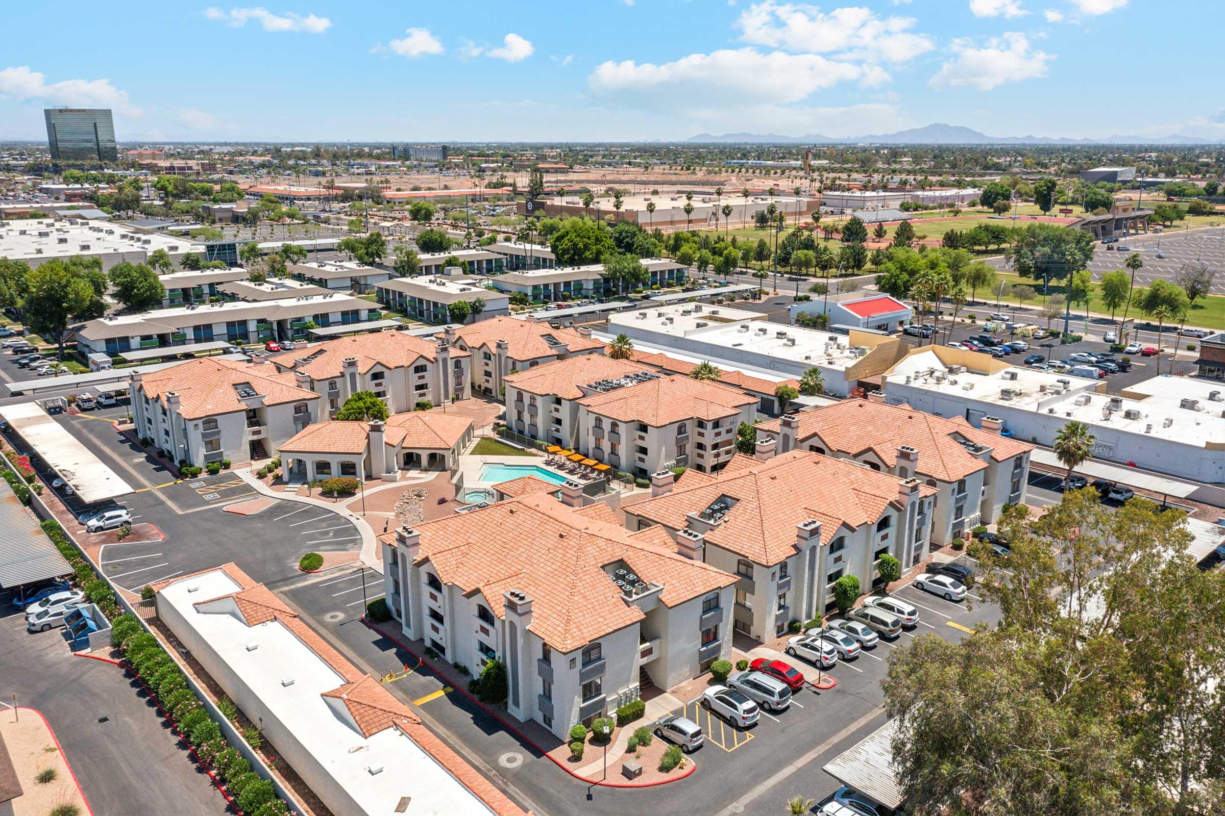 Aerial view of a residential complex featuring multiple buildings with terracotta roofs. The area includes a swimming pool and landscaped grounds, surrounded by parking lots. In the background, there are additional commercial buildings and open spaces under a clear blue sky.