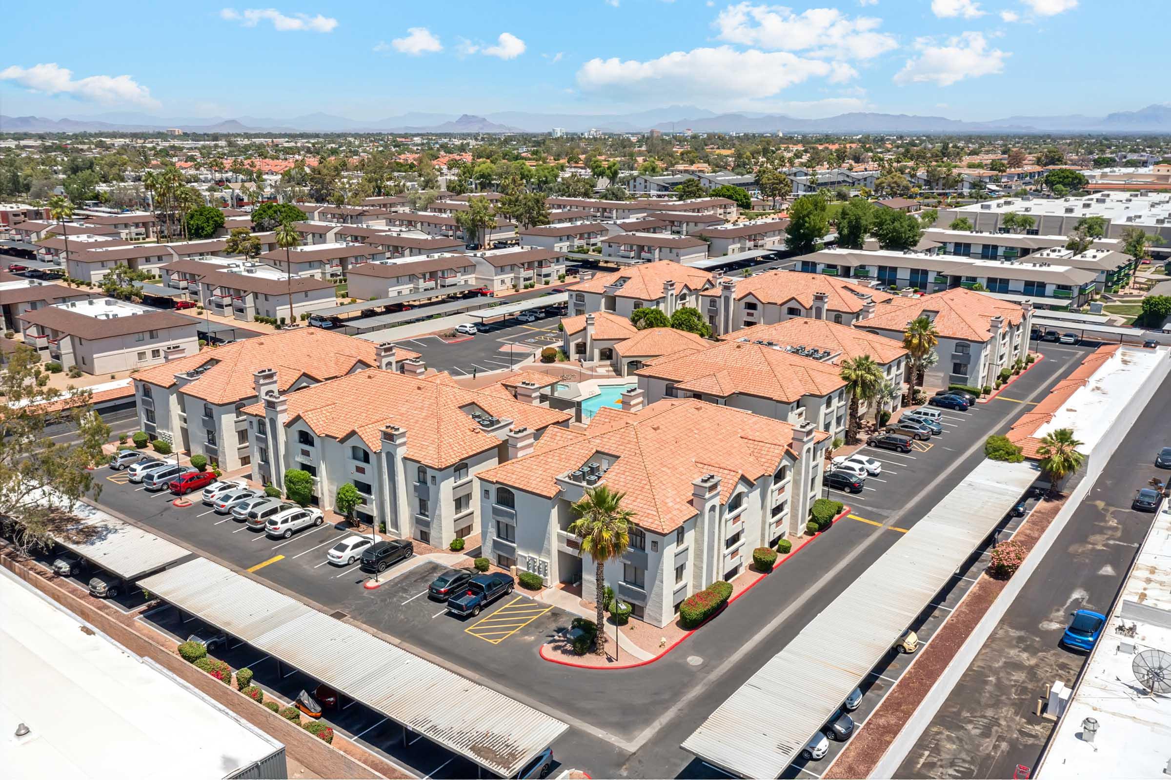Aerial view of an apartment complex featuring several buildings with terracotta roofs, surrounded by parking spaces and palm trees. A swimming pool is visible in the central area. The background shows more residential buildings and hills under a clear sky.
