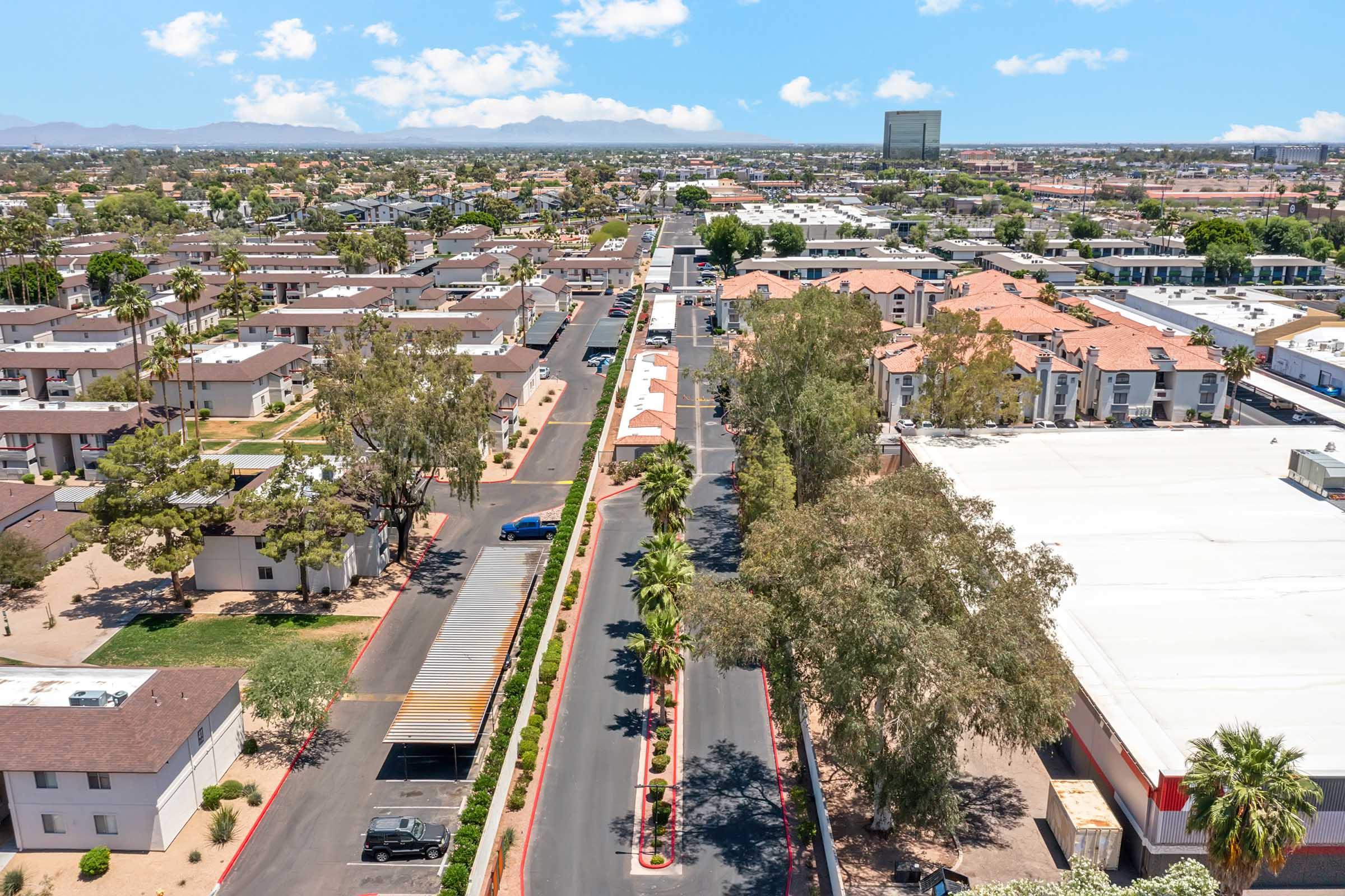 Aerial view of a suburban neighborhood featuring rows of houses, palm trees, and a wide street lined with parked cars. In the background, commercial buildings and a distant mountain range are visible under a clear blue sky with scattered clouds.