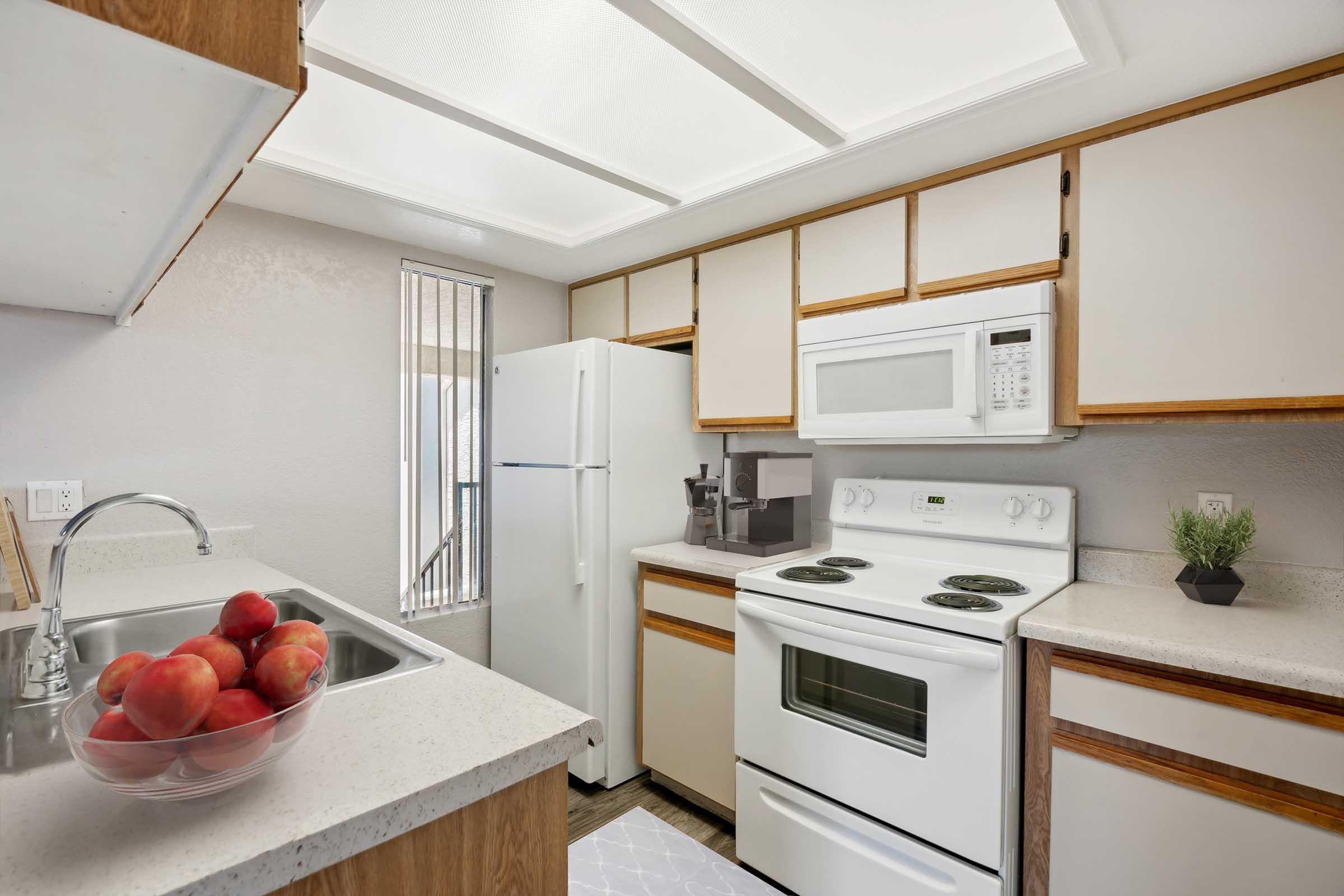A modern kitchen featuring white cabinetry, a countertop with a fruit bowl filled with red apples, a stainless steel sink, a refrigerator, a microwave, and an electric stove. Soft lighting illuminates the space, complemented by a light-colored floor.